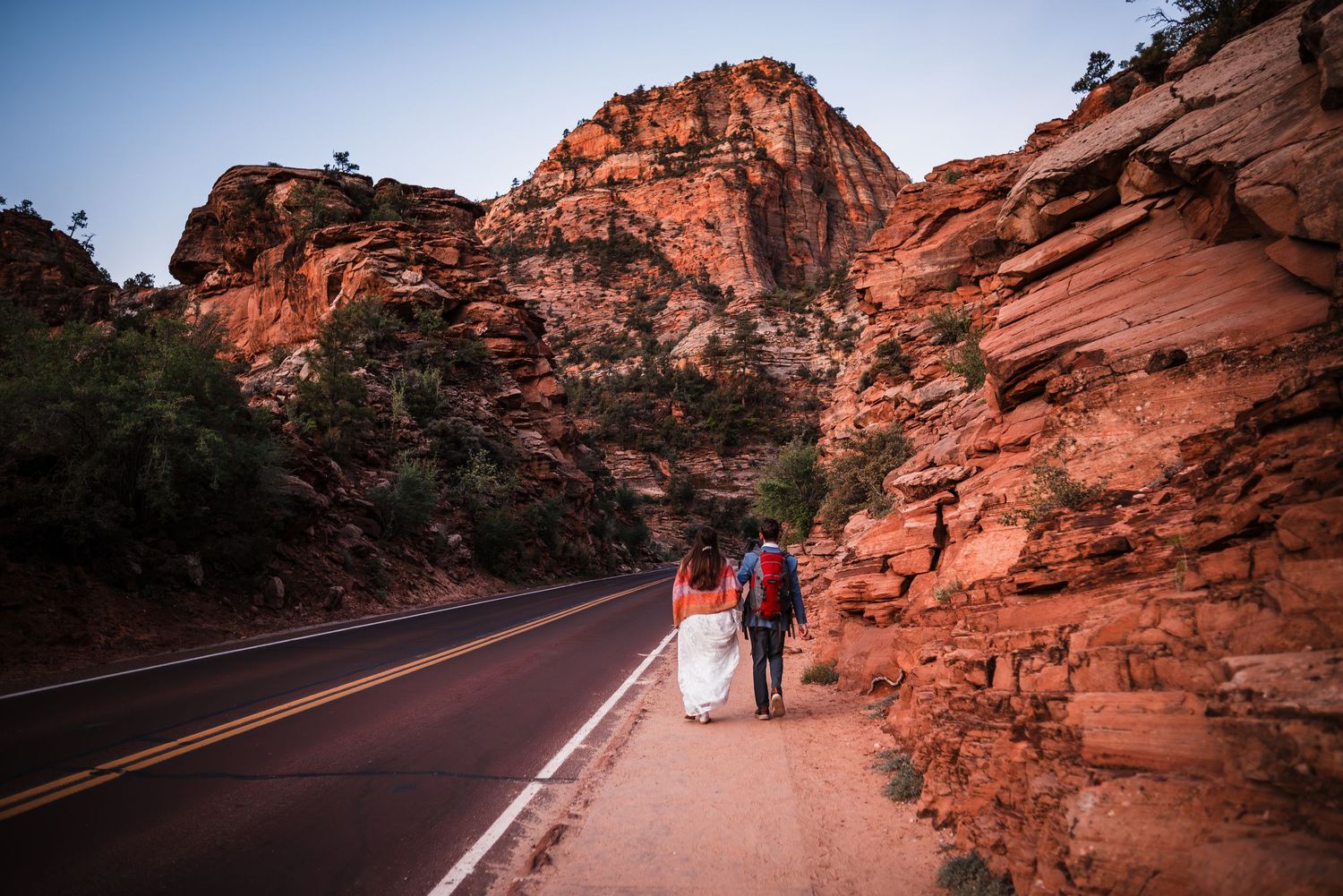 Bride and groom walking along a scenic red rock canyon road at Zion National Park during golden hour.