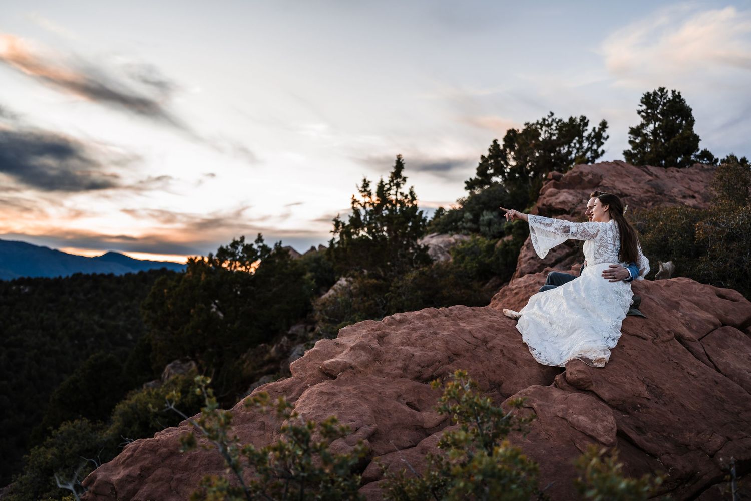 Bride in lace wedding dress sitting on red rocks at sunset with mountain views at Garden of the Gods.