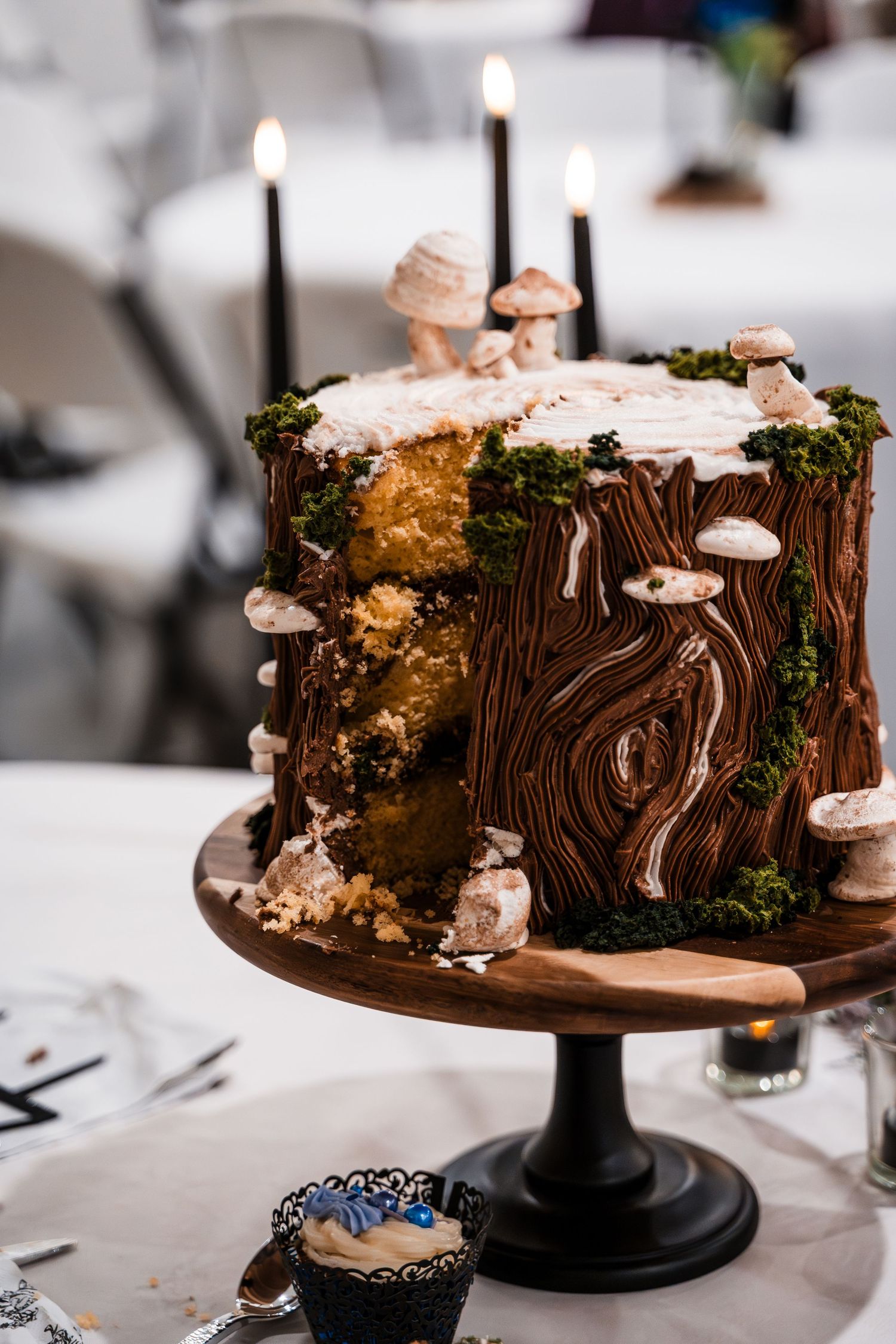 Elegant tree stump wedding cake with mushroom decorations, moss, and black candles on wooden stand.