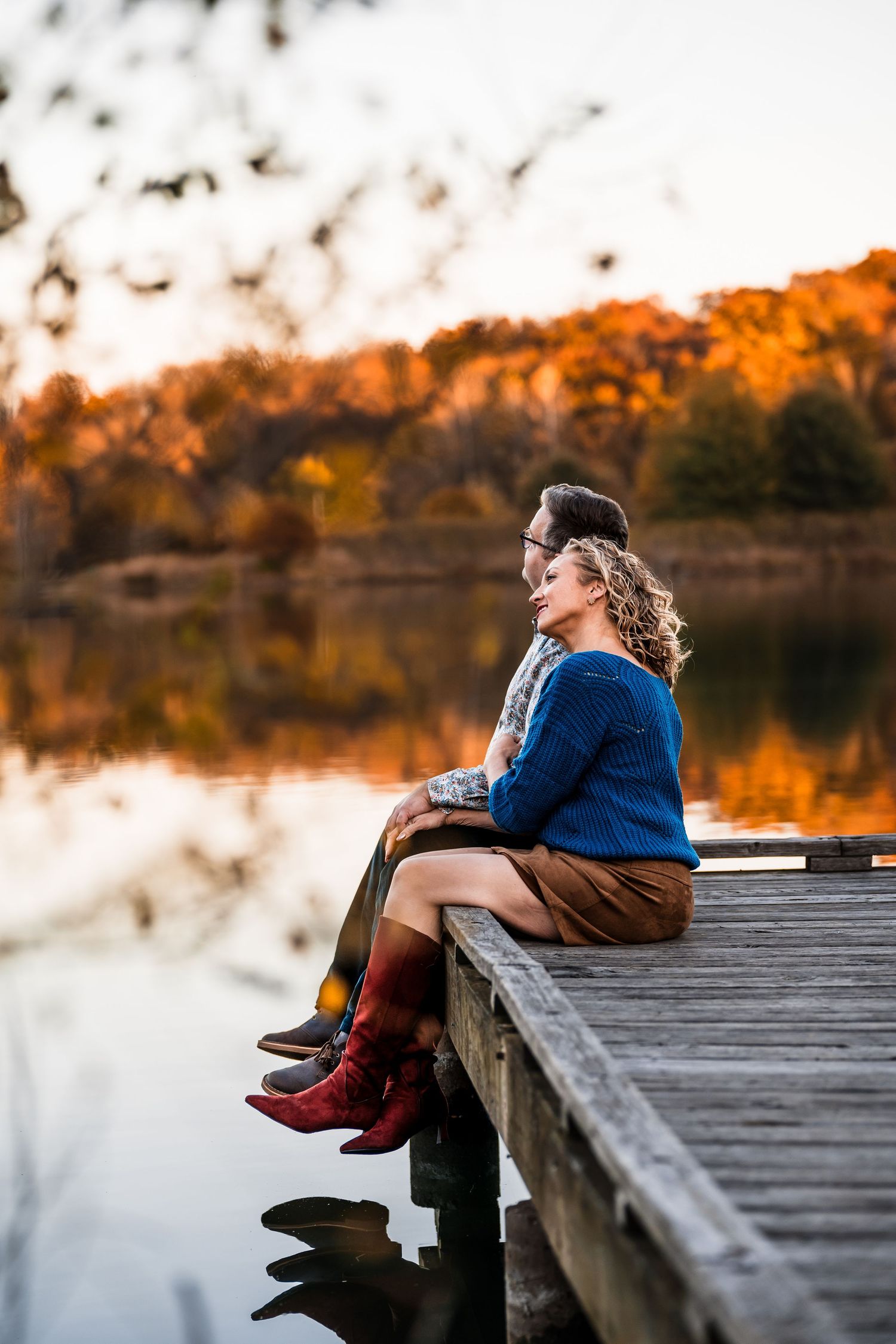 Someone sits on a wooden dock by a calm lake during golden hour sunset with autumn colors reflecting in the water.