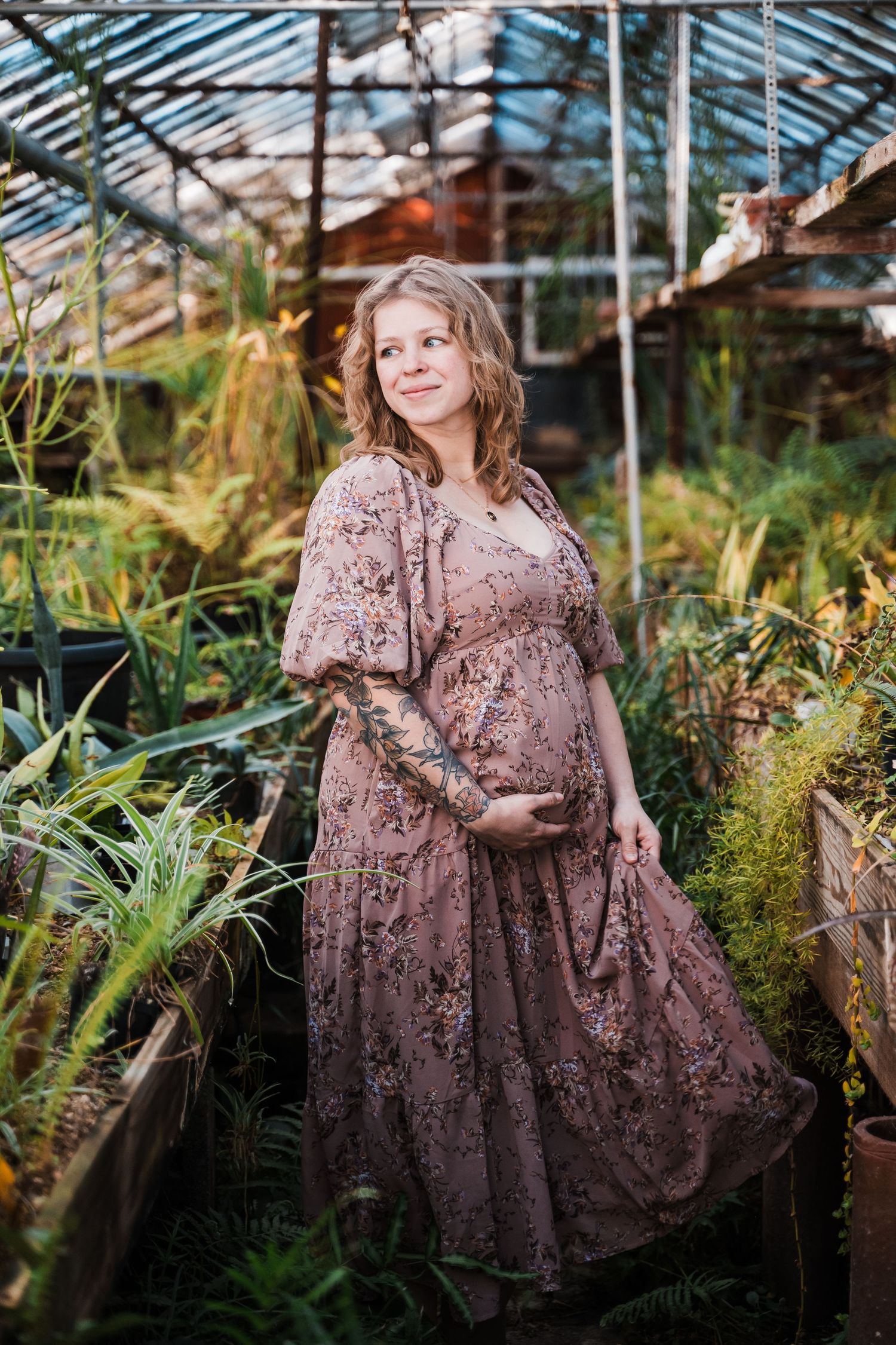 A person in a brown floral dress poses gracefully among lush ferns and greenery in a greenhouse setting.