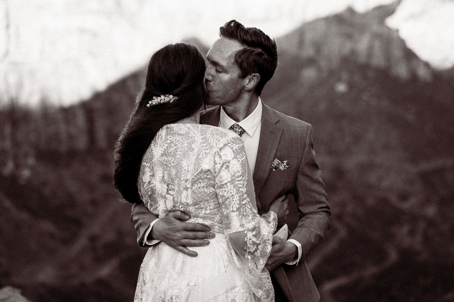 Bride and groom share a romantic kiss with mountain backdrop in black and white wedding portrait.