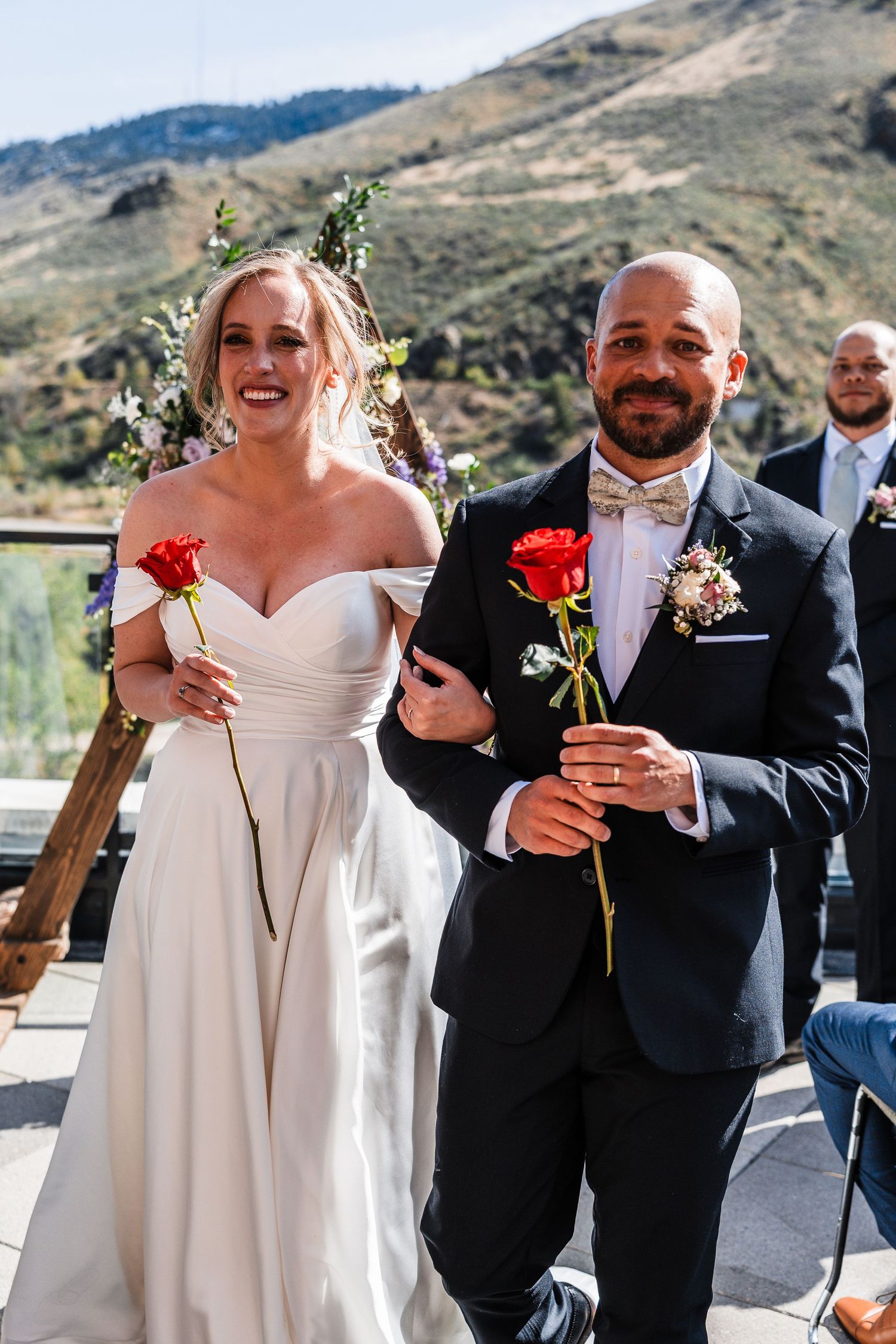 Bride and groom walk down the aisle holding red roses at an outdoor mountain wedding ceremony.