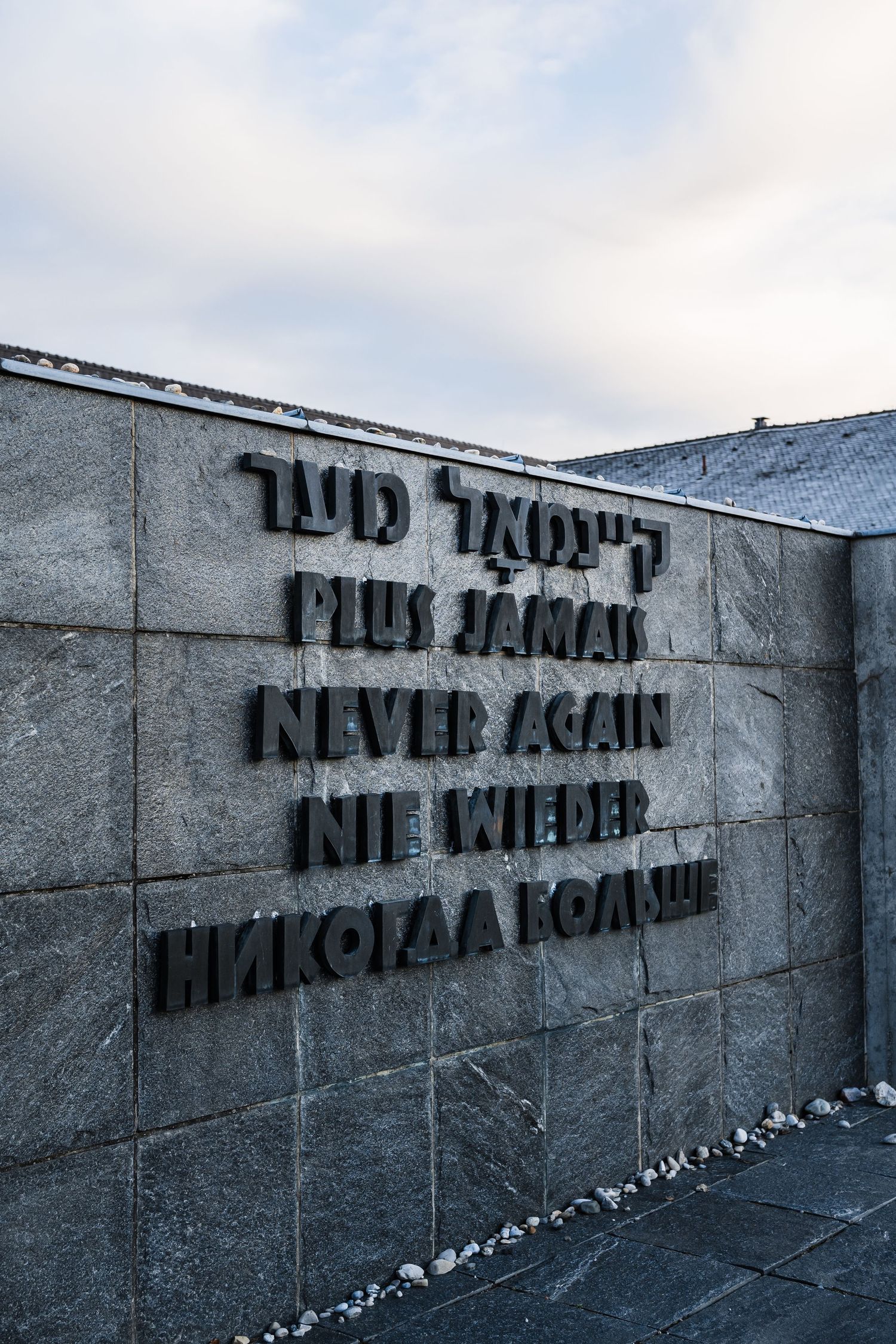 Memorial wall with 'Never Again' text painted in black letters on concrete surface in multiple languages.