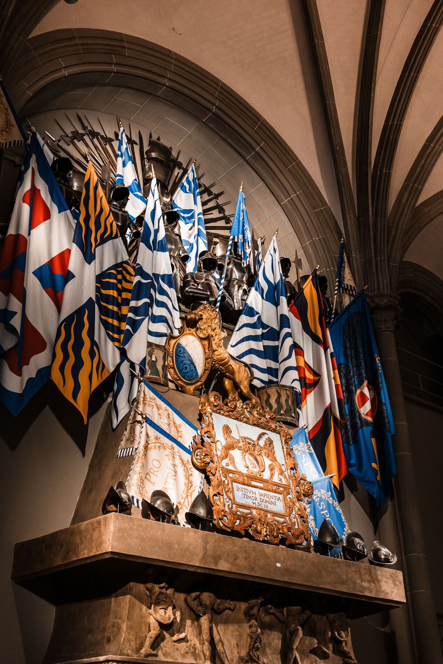 Ornate medieval heraldic display with blue and white flags adorning a stone wall under gothic arches.