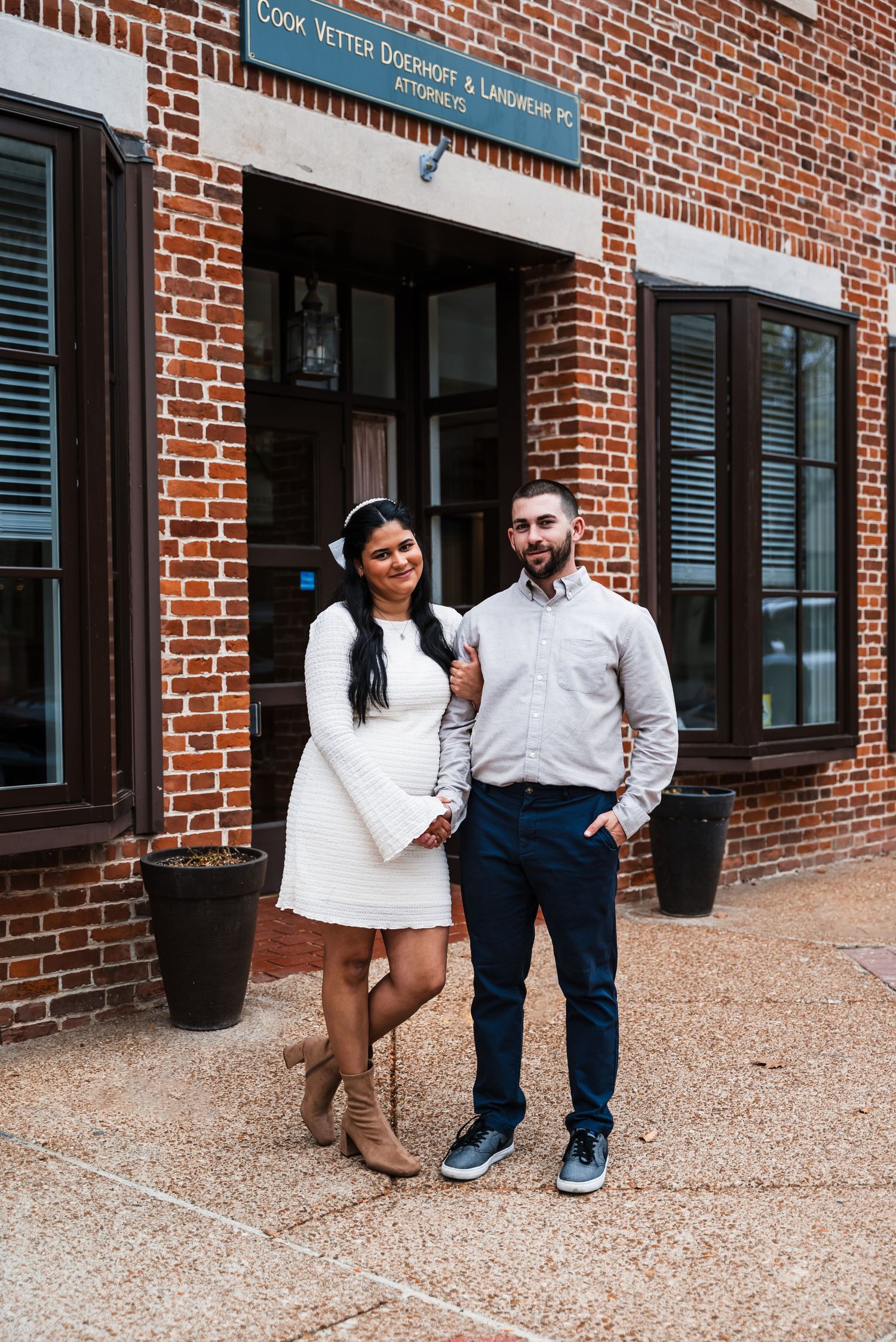 A couple poses in front of a brick building entrance with black shuttered windows.