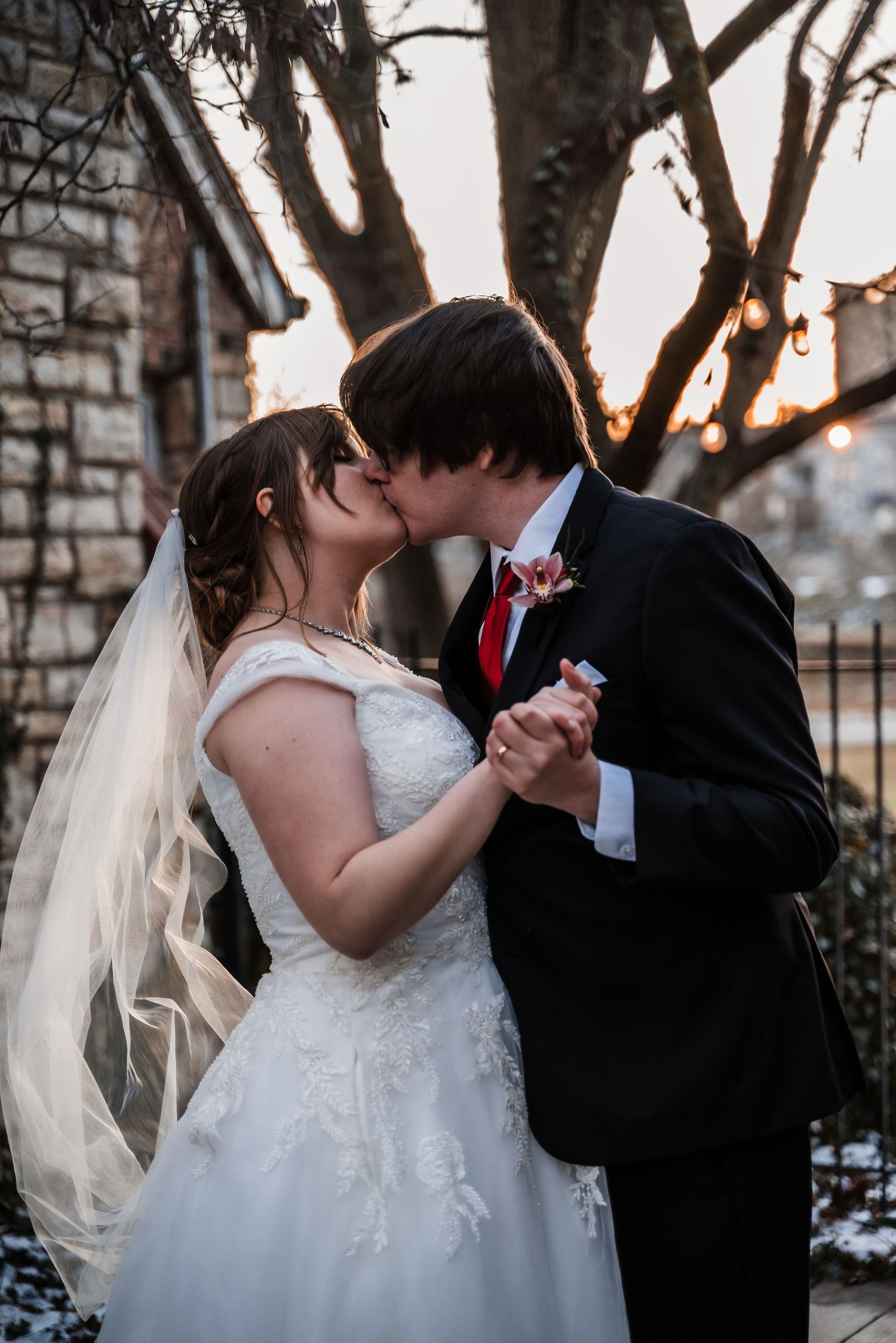 A romantic kiss between newlyweds during their winter wedding with string lights and trees in the background.