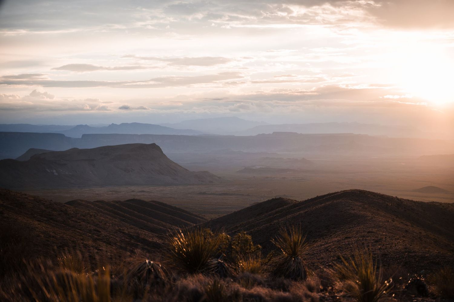Dramatic sunset over desert mountain ranges with golden light illuminating the rugged landscape.