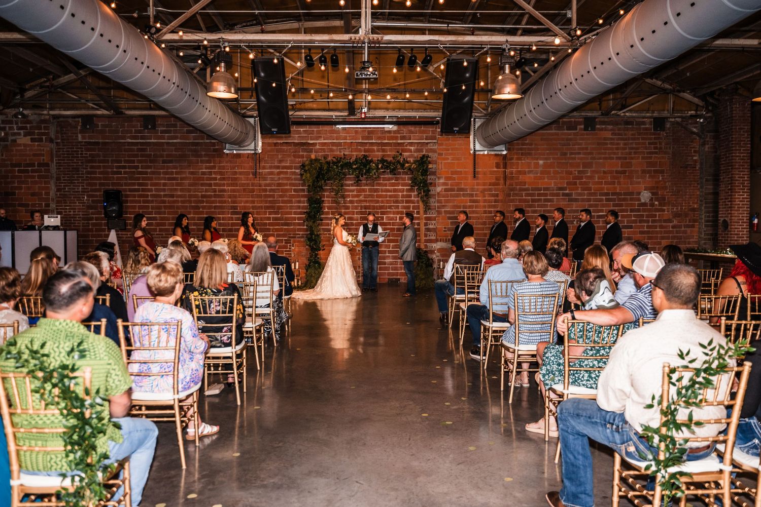 Wedding ceremony in an industrial brick venue with exposed ductwork and string lights overhead.
