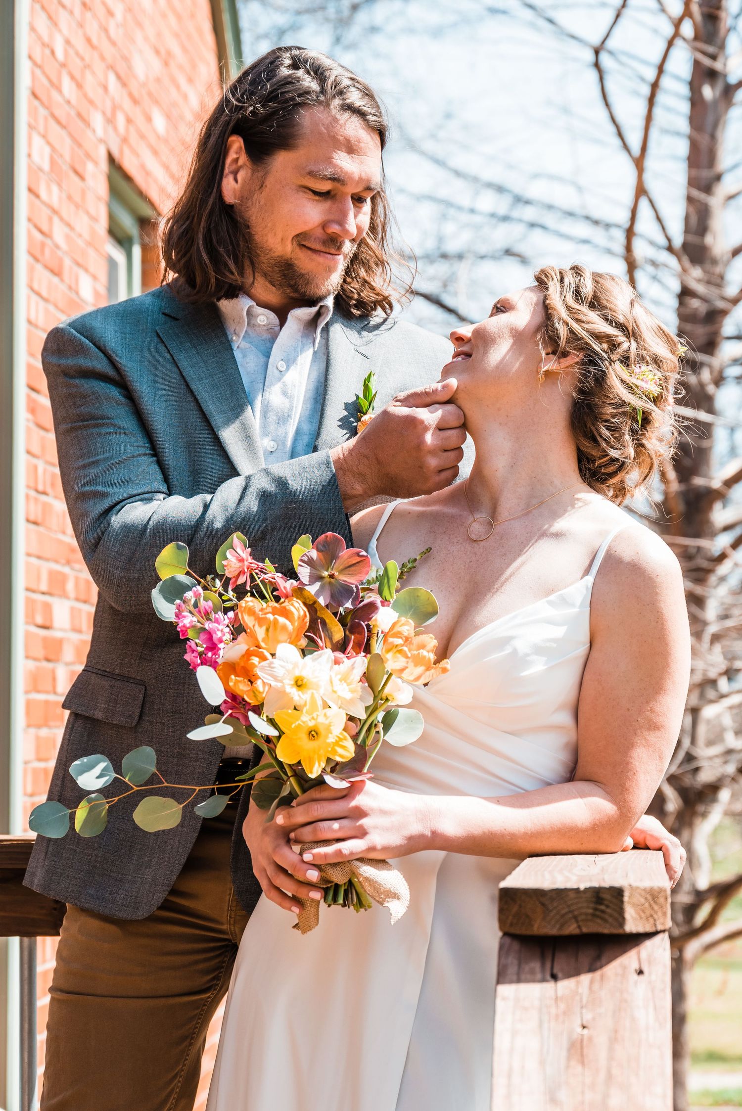 Newlyweds share intimate moment with vibrant spring bouquet against brick building.