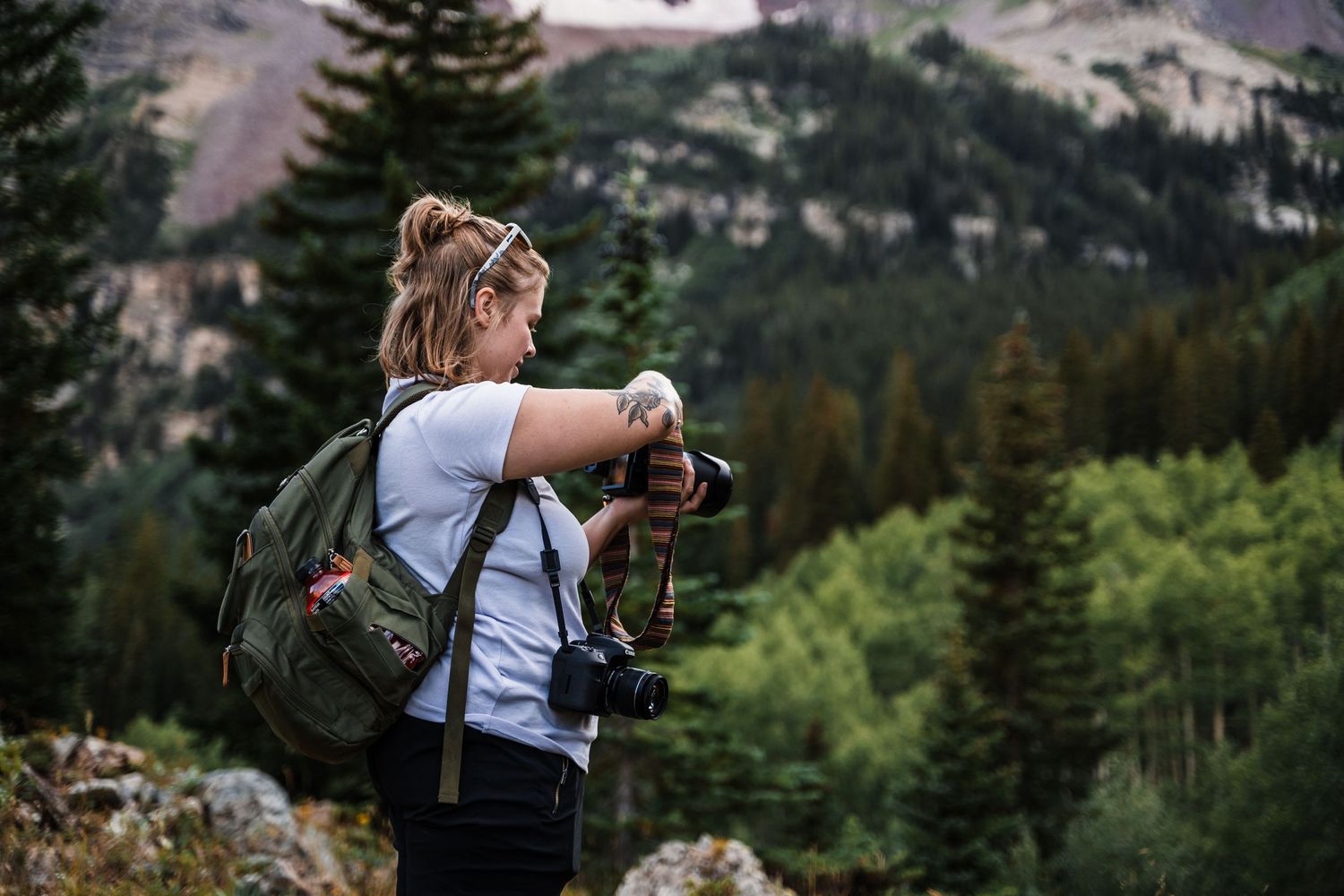 A photographer with a backpack takes photos in a mountainous landscape surrounded by evergreen trees and peaks.