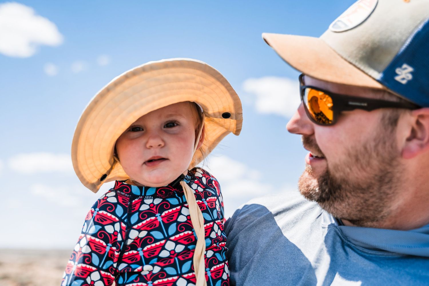 A child wearing a tan cowboy hat and colorful outfit sits next to someone in sunglasses against a blue sky background.