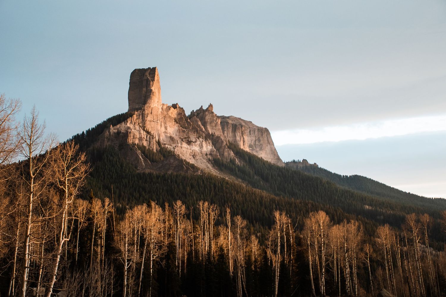 A dramatic rock formation towers above winter aspen trees at sunset in Colorado's wilderness.