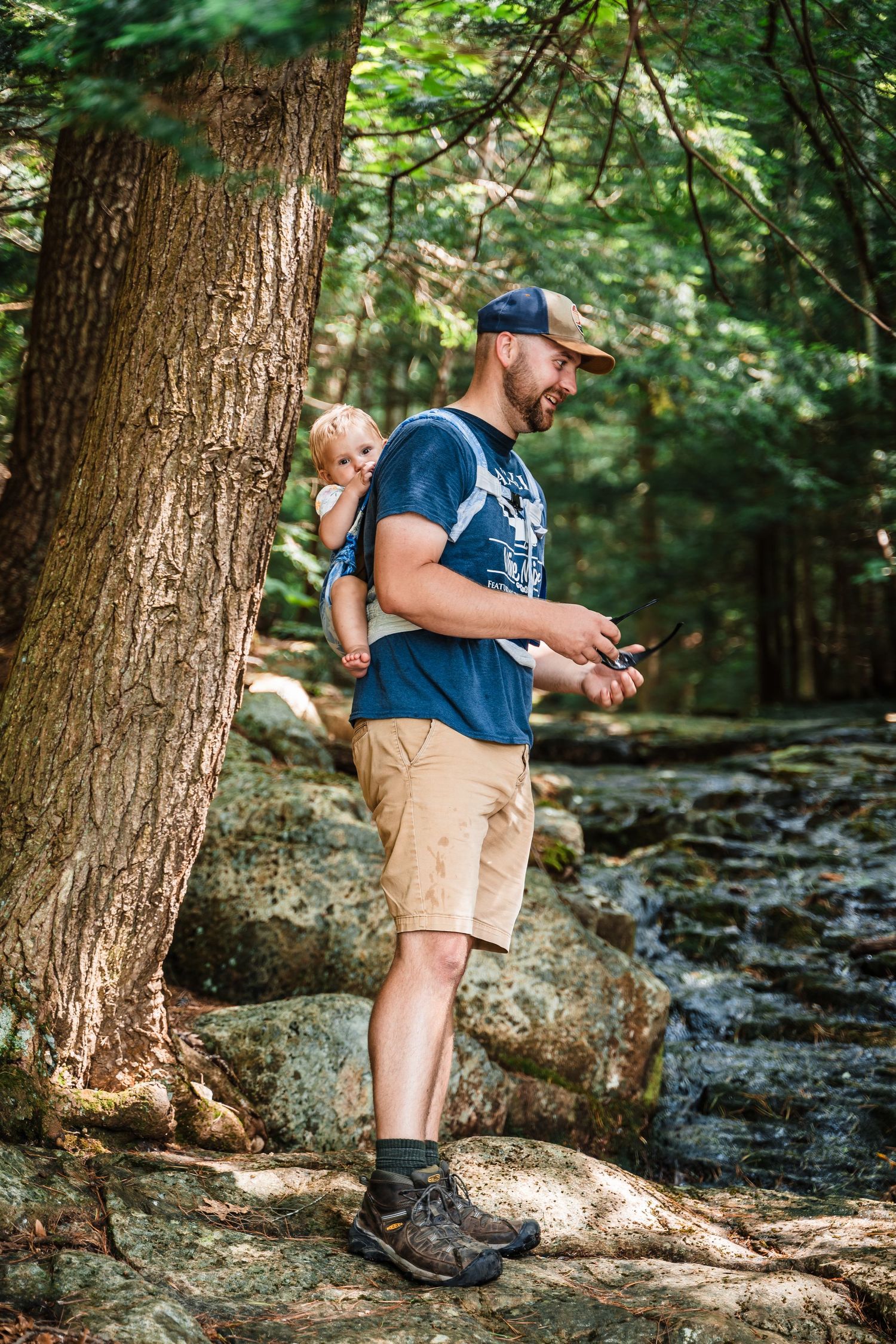 Outdoor hiking scene along a rocky stream in a lush forest with hiker wearing blue shirt and khaki shorts observing nature.