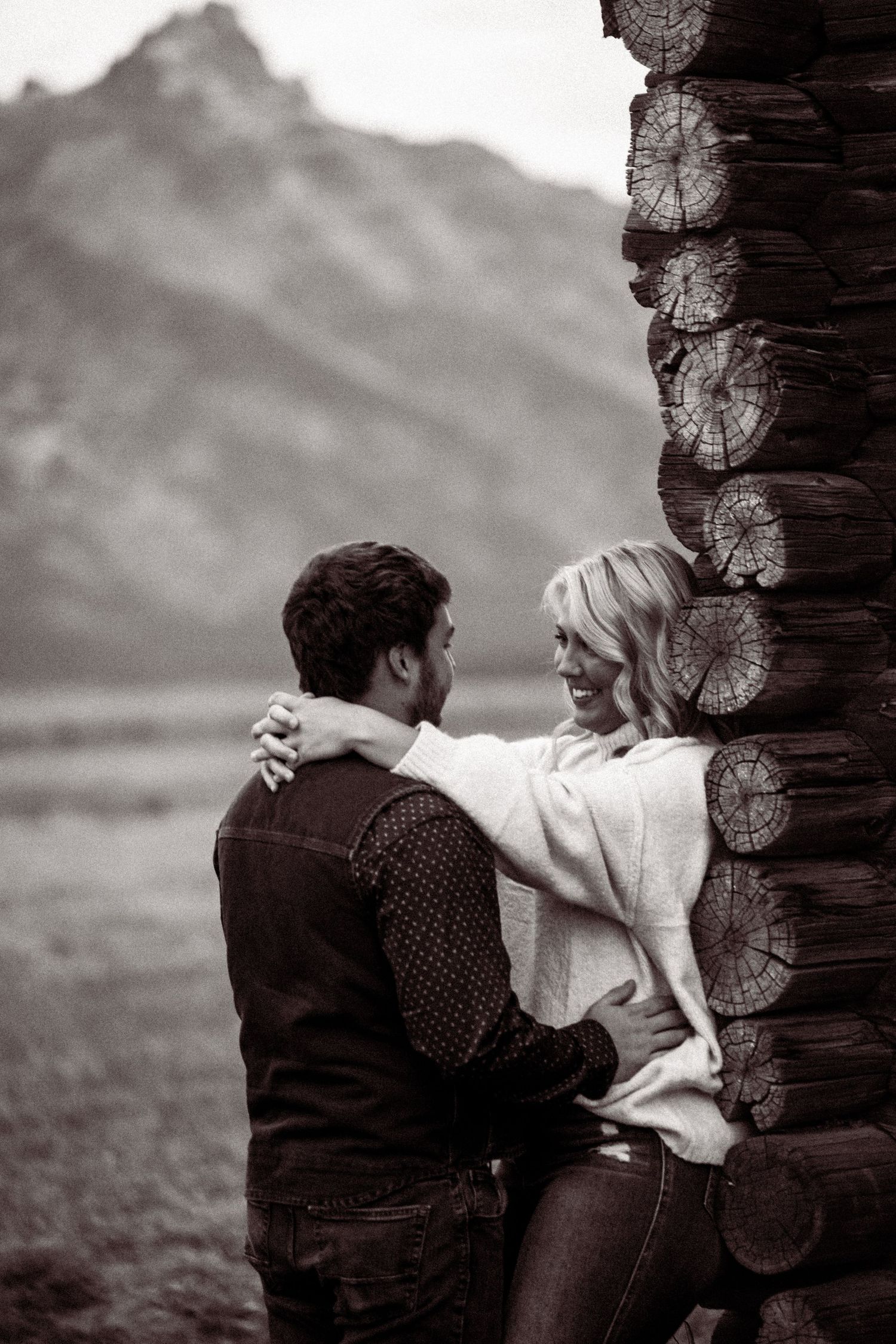 Black and white photo of a couple sharing an intimate moment against a rustic log cabin wall.