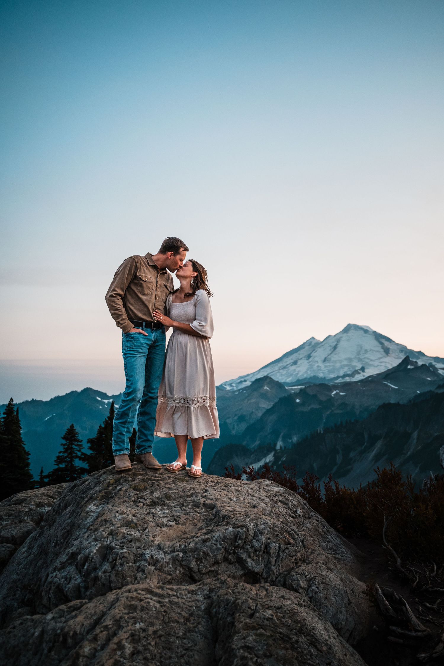 Couple sharing a romantic moment on a rocky outcrop with snow-capped mountain peaks in the background at dusk.