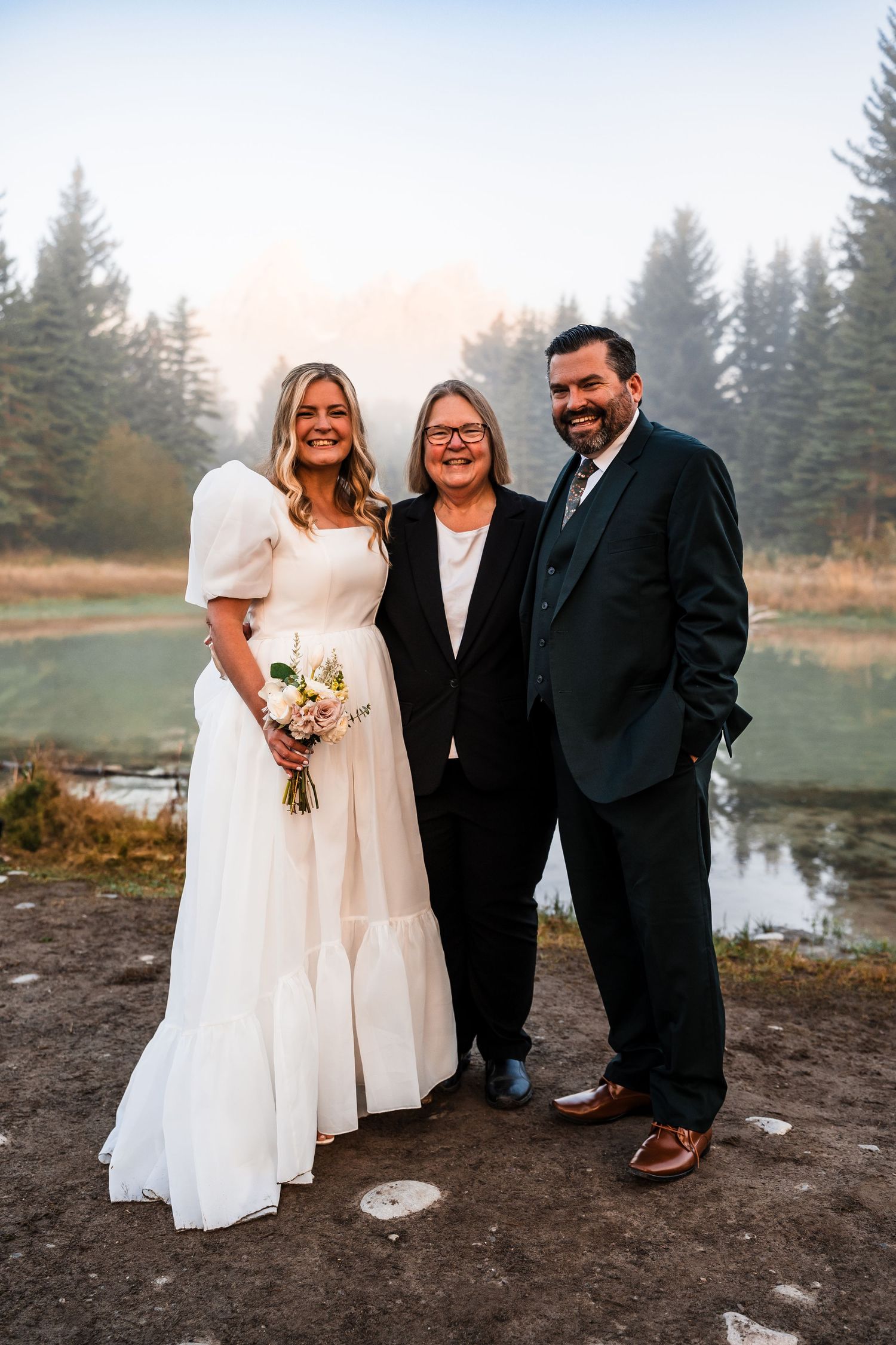 Wedding party poses together by a lake with mountains and evergreen trees in the background.