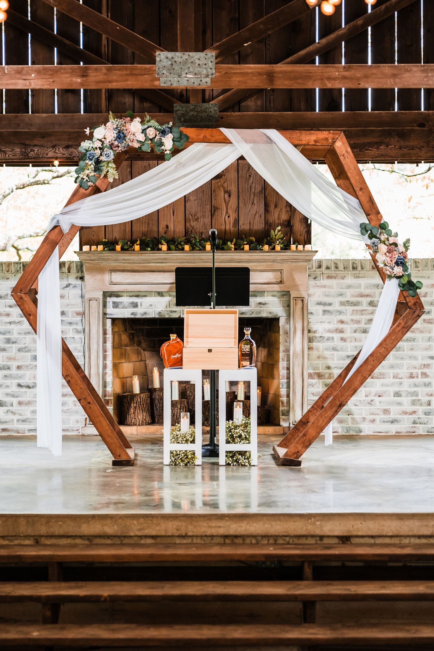 Modern hexagonal wedding arch decorated with white fabric and floral arrangements inside a rustic barn venue.