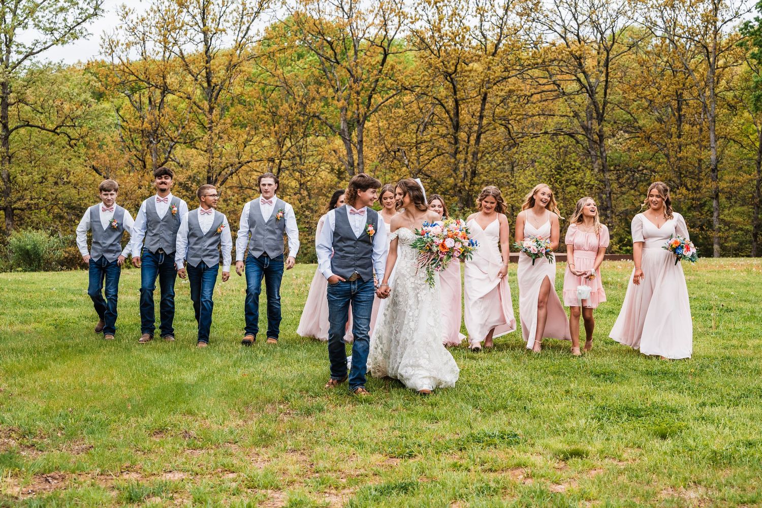 Wedding party in formal attire poses together on a grassy field surrounded by autumn trees.