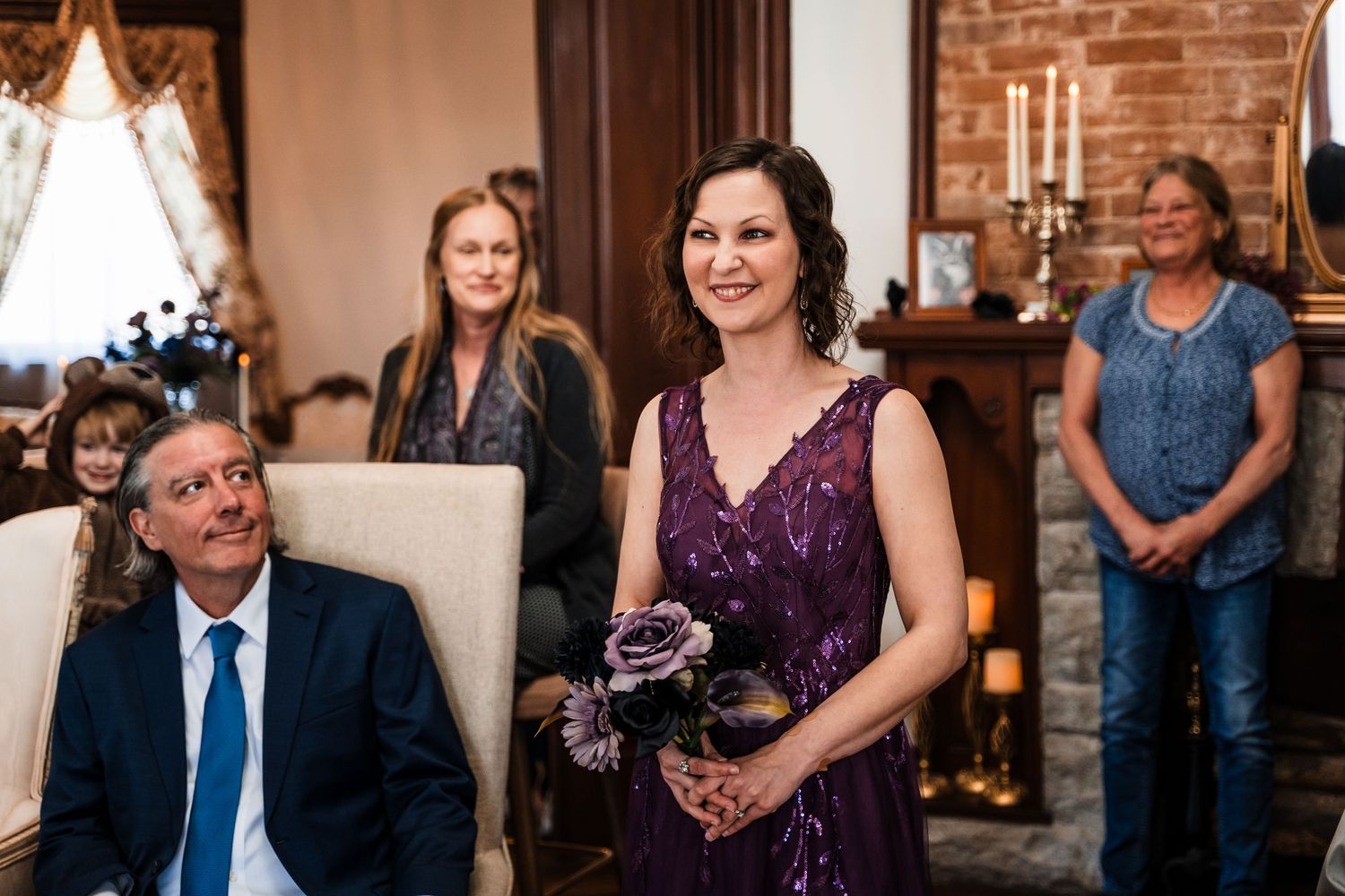 Smiling bridesmaid in purple gown holds bouquet during intimate indoor wedding ceremony.