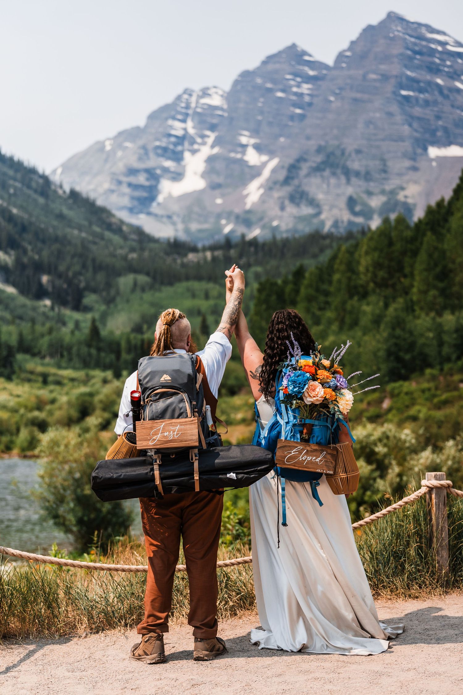 Newlyweds raise hands holding Just Eloped signs on backpacks before iconic Maroon Bells mountains.