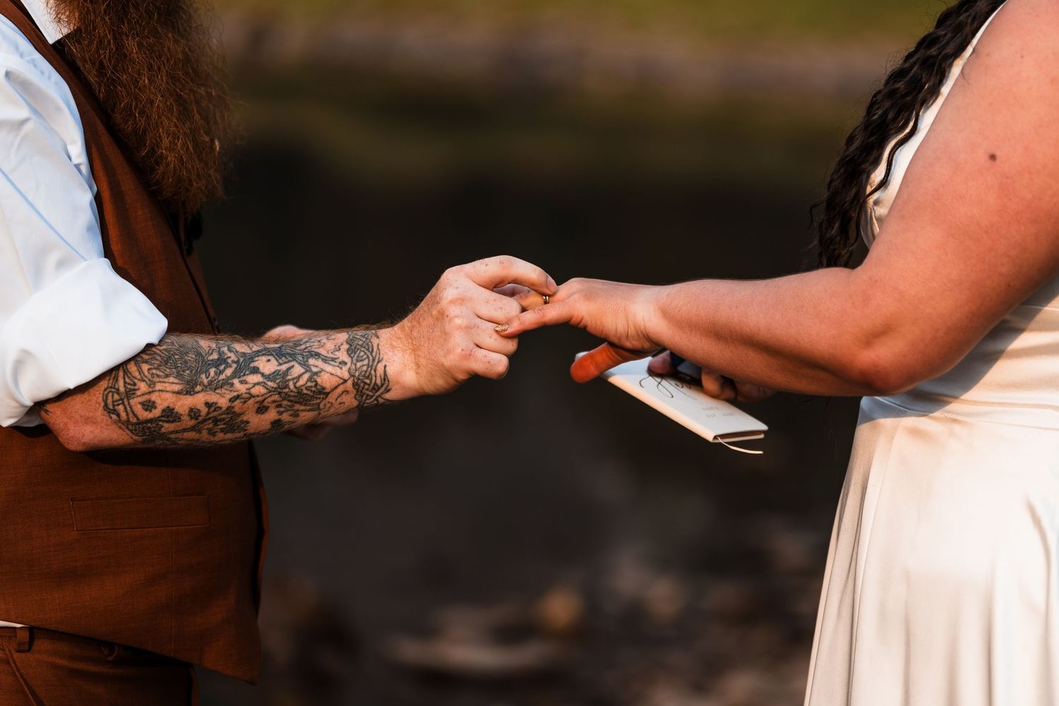 Close-up of tattooed groom placing a ring on brides finger during an intimate outdoor elopement ceremony.