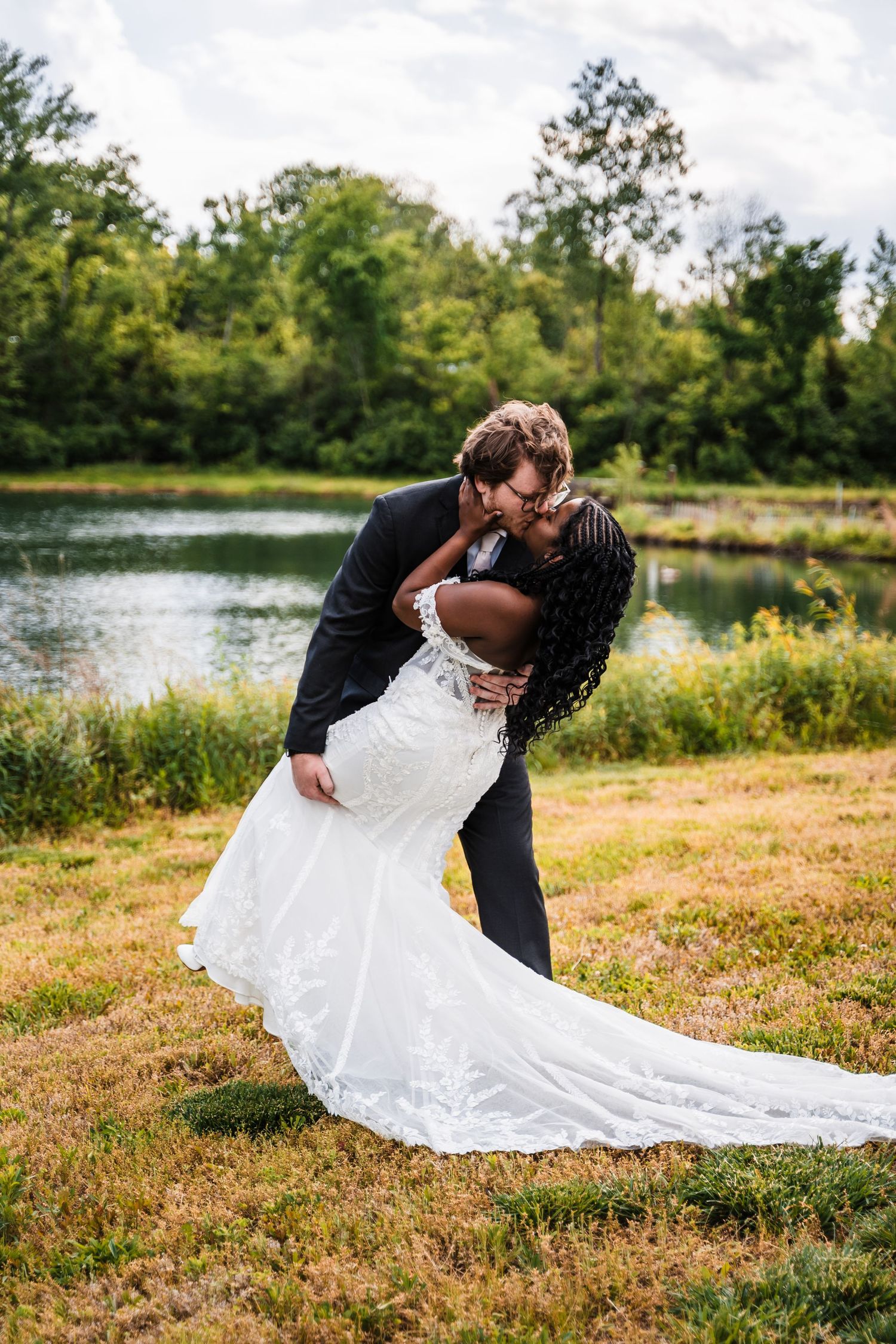Interracial couple sharing a romantic dip kiss in front of a serene pond, bride in lace mermaid gown.