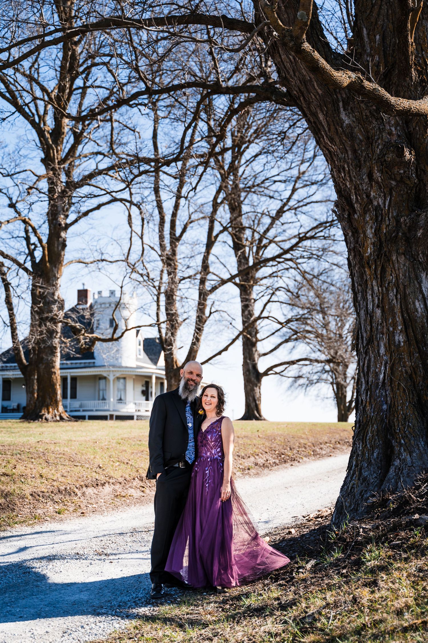 Couple posing by large bare trees on gravel path with white farmhouse in background on sunny winter day.