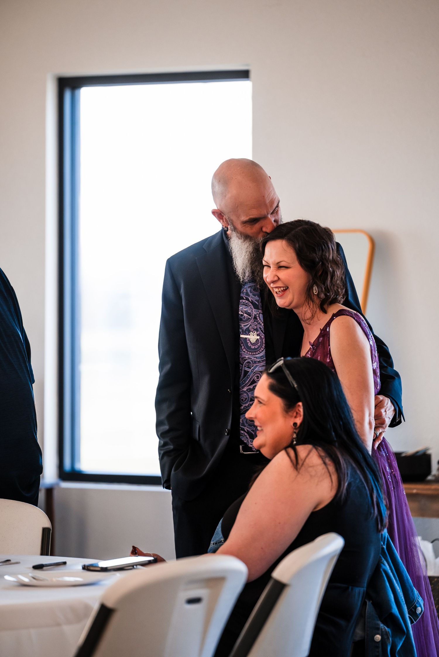 Bald bearded groom in black suit laughing with bride in purple dress at wedding reception.