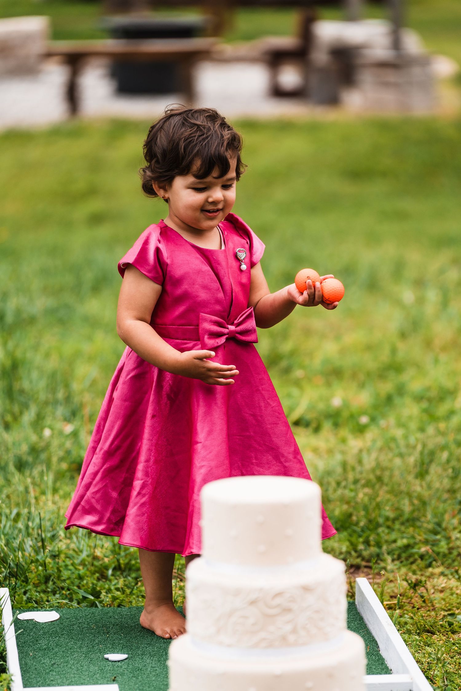 Toddler in pink dress playing with golf balls near white wedding cake at outdoor reception.