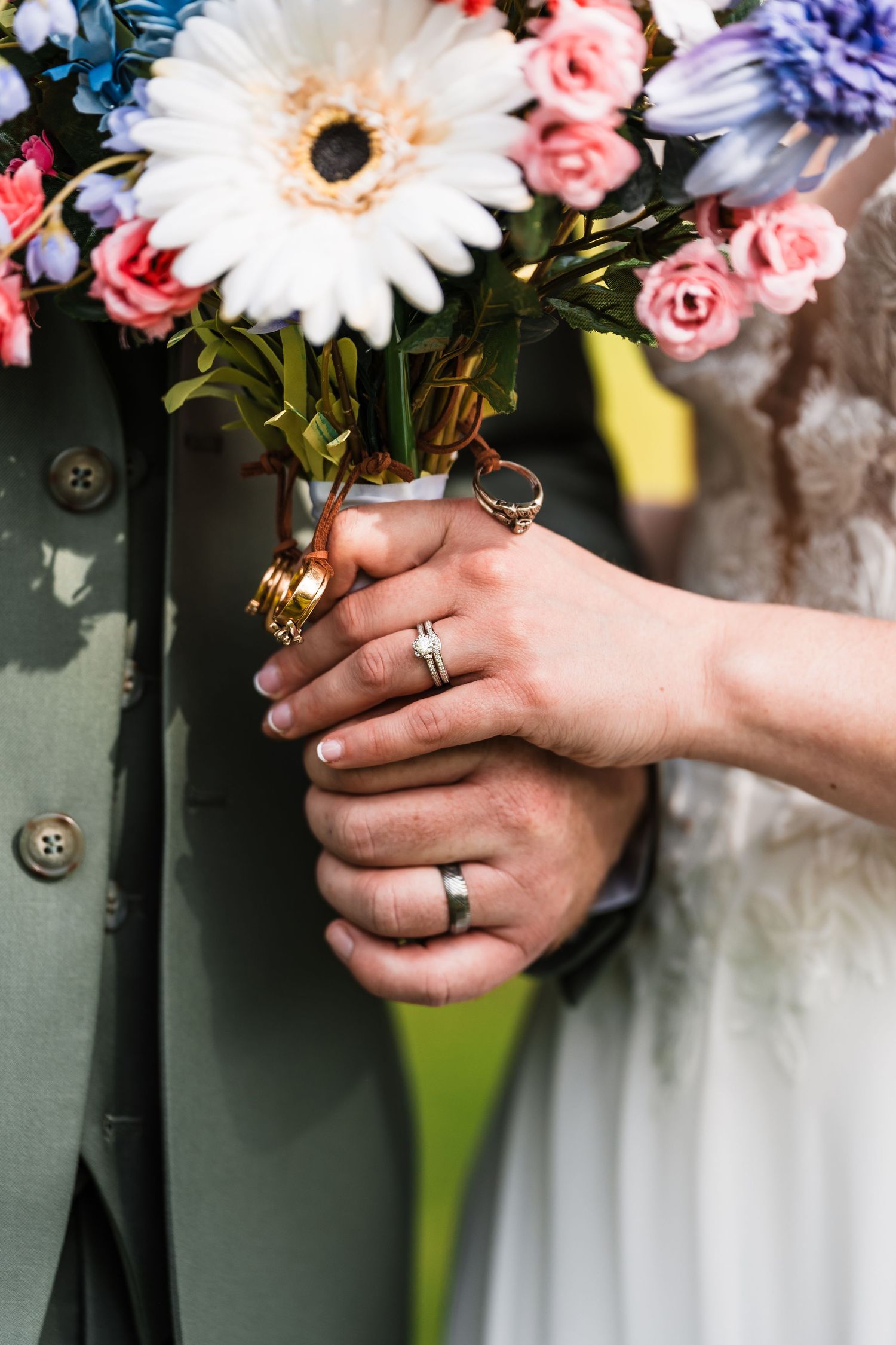 Close-up of bride and groom hands holding colorful floral bouquet, wearing wedding rings and bands.