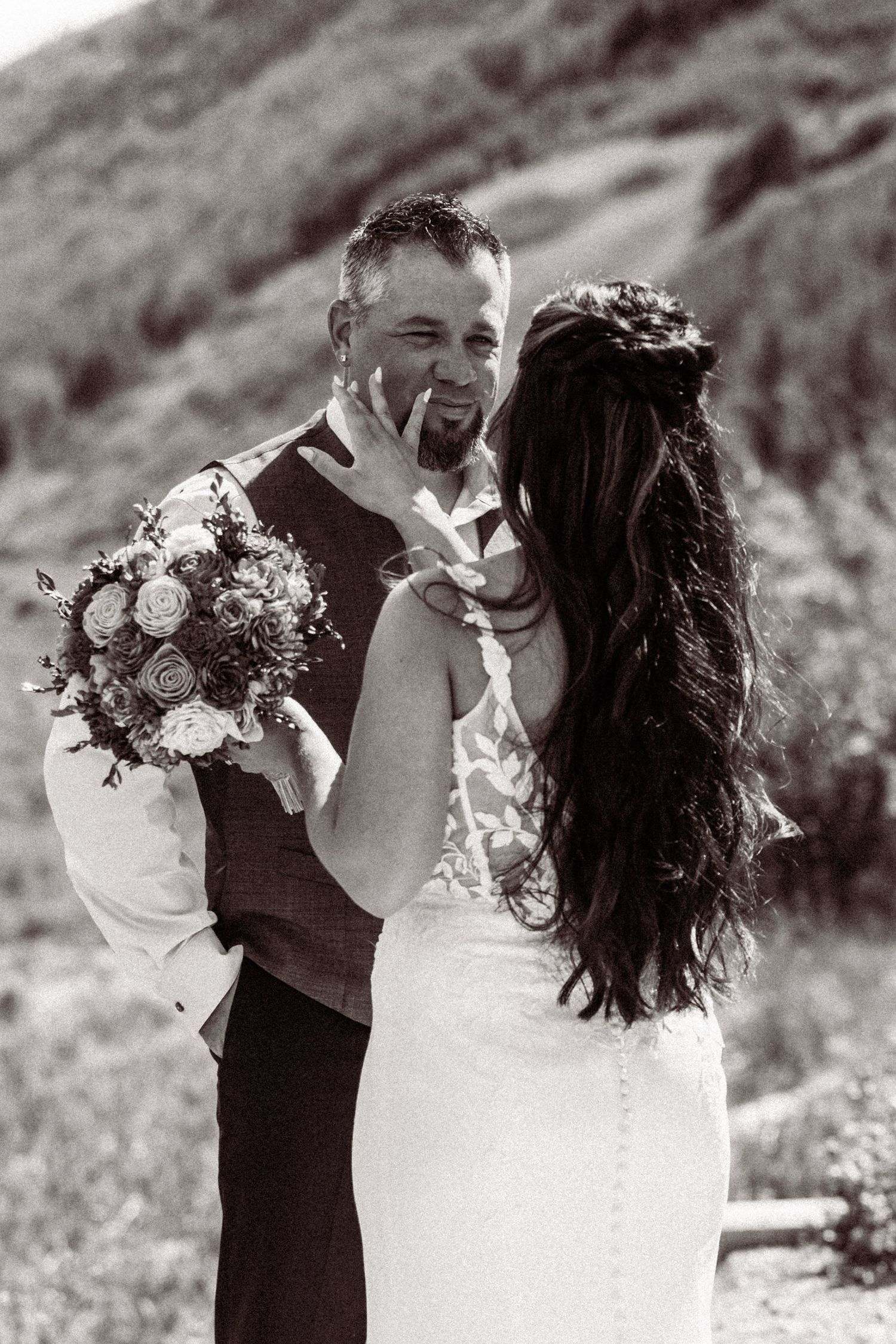 Black and white photo of a groom kissing bride's hand while she holds a floral bouquet outdoors.