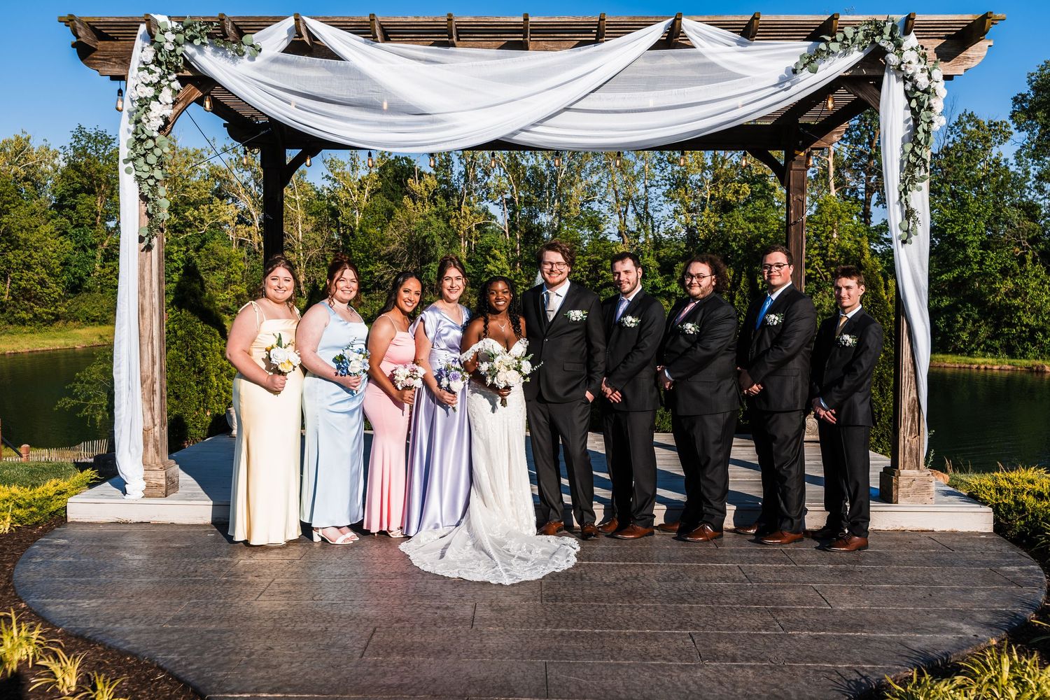 Wedding party poses under floral draped pergola outdoors, bridesmaids in pastel gowns and groomsmen in dark suits.