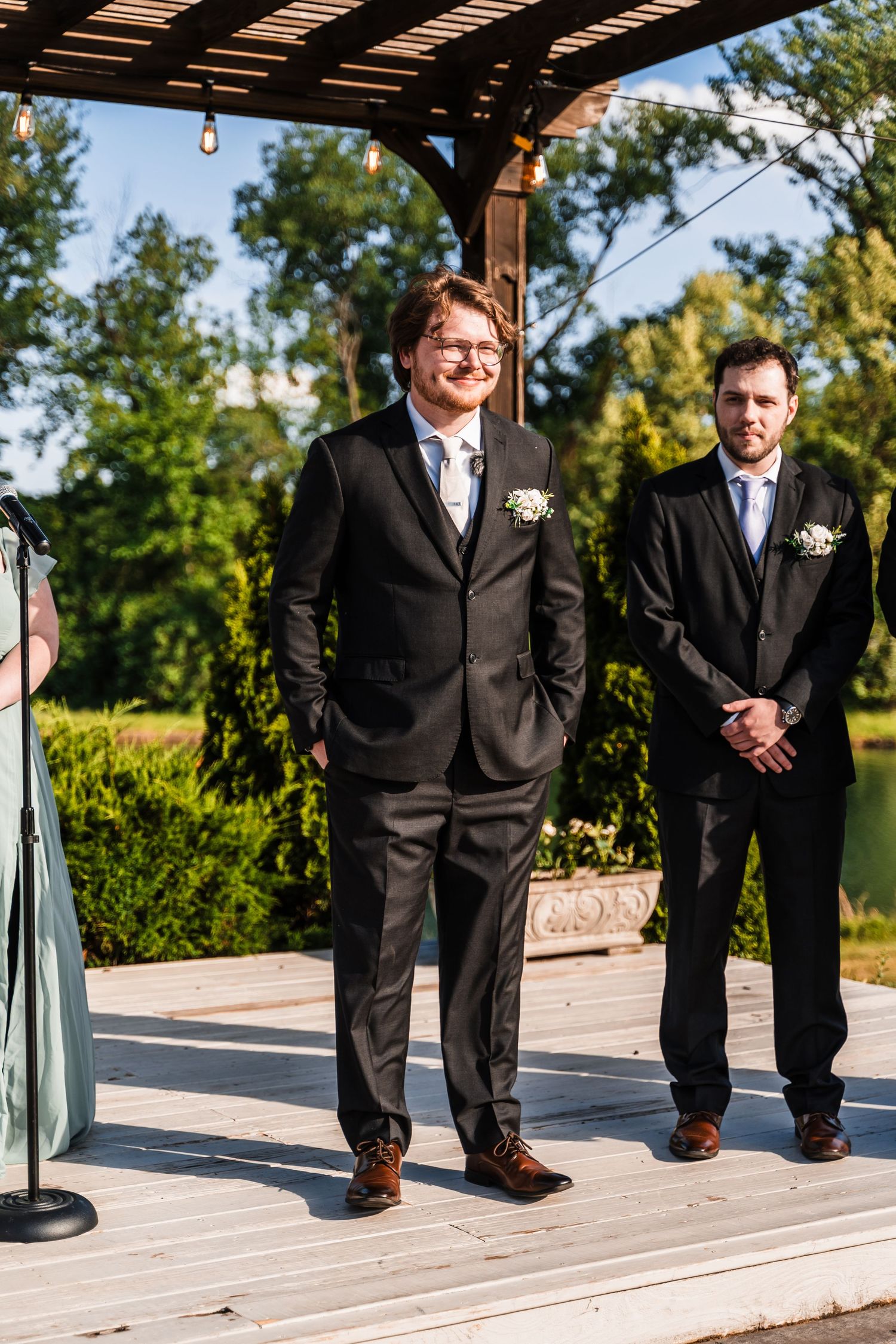 Groom in black suit smiles at altar with groomsman behind him at outdoor wedding ceremony.