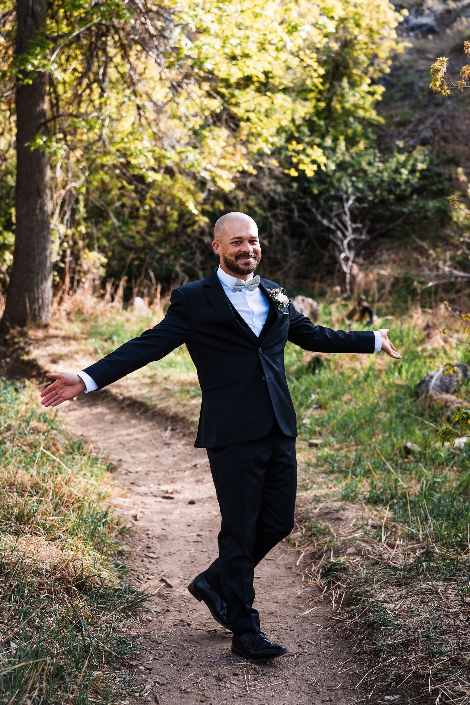 Smiling bald groom in black tuxedo with boutonniere poses with arms outstretched on a forest path.