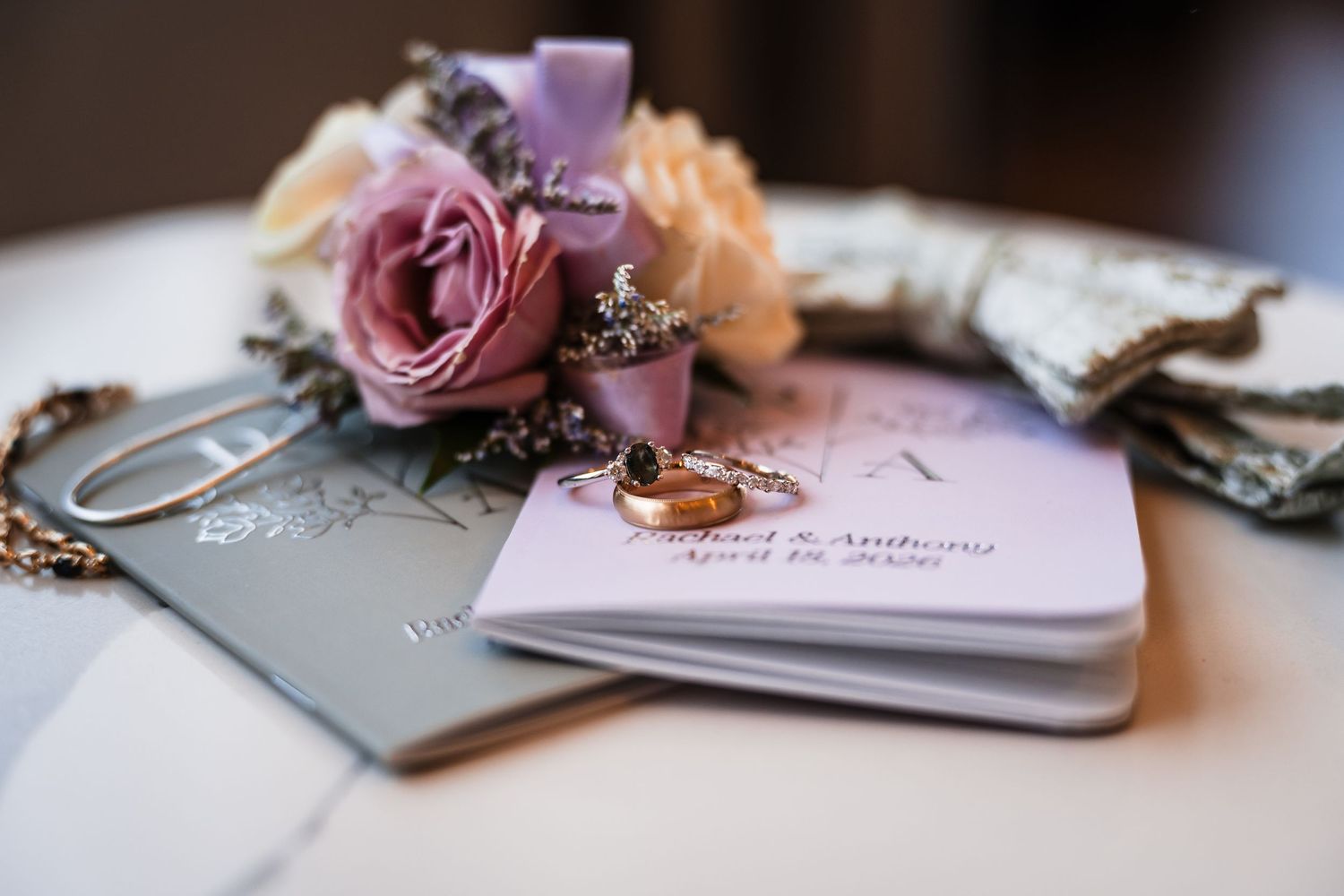 Wedding rings and corsage rest atop personalized ceremony booklets on a white table.