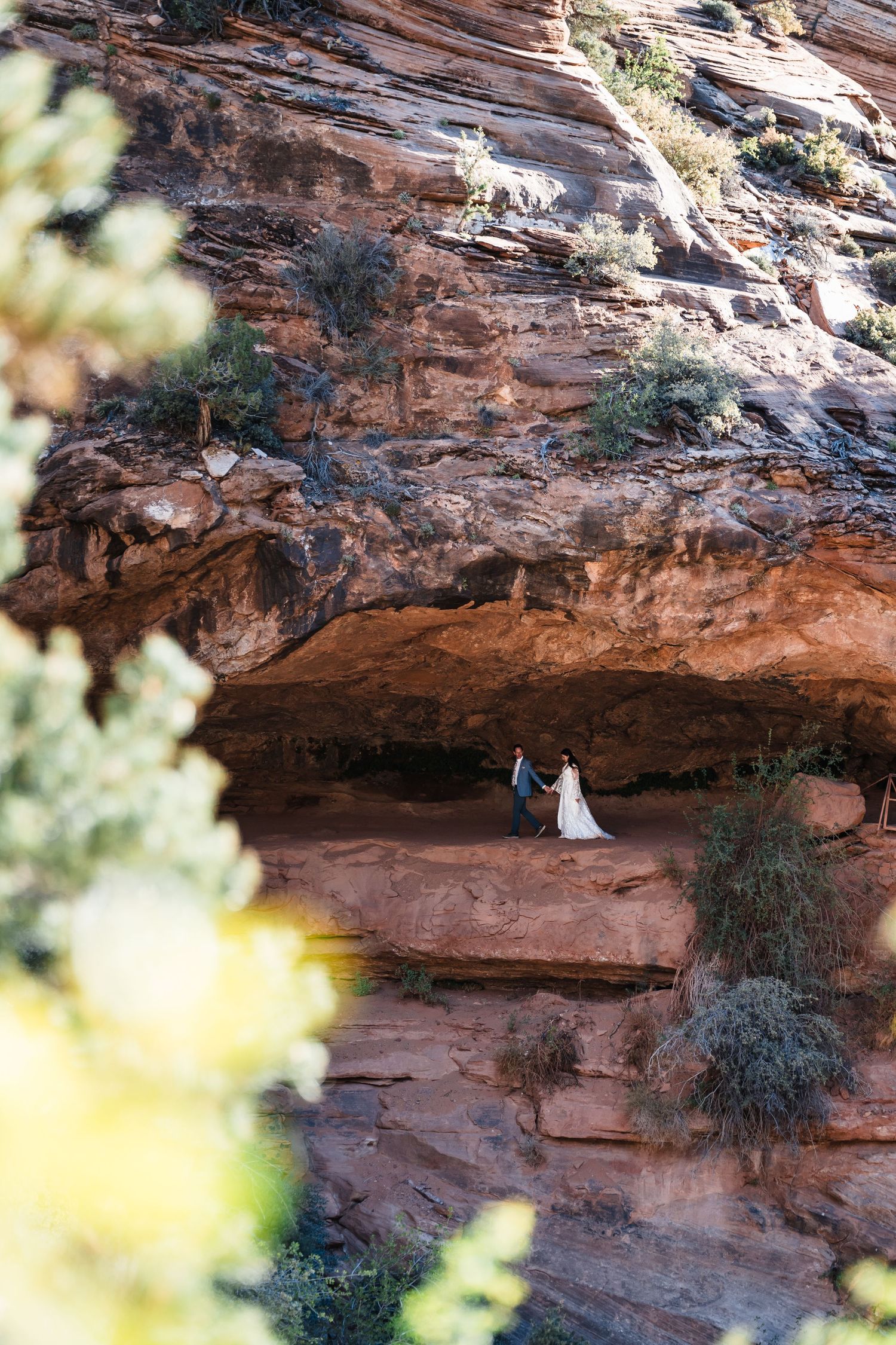Couple in wedding attire standing beneath a dramatic red rock canyon overhang in a desert landscape.