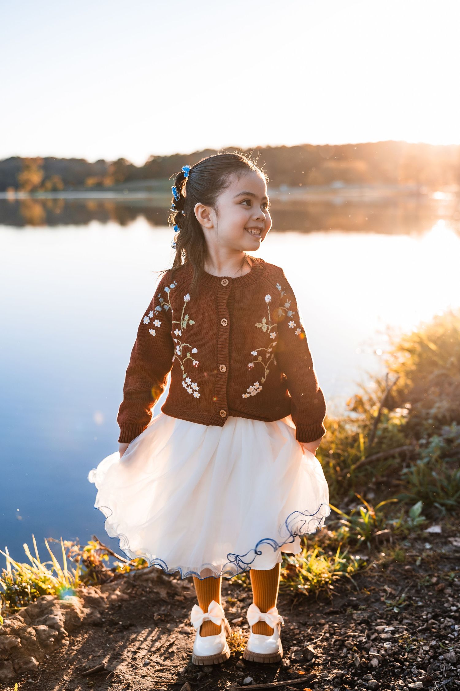 A child in a white tulle dress and dark cardigan plays by a lake at sunset, creating a dreamy outdoor portrait.