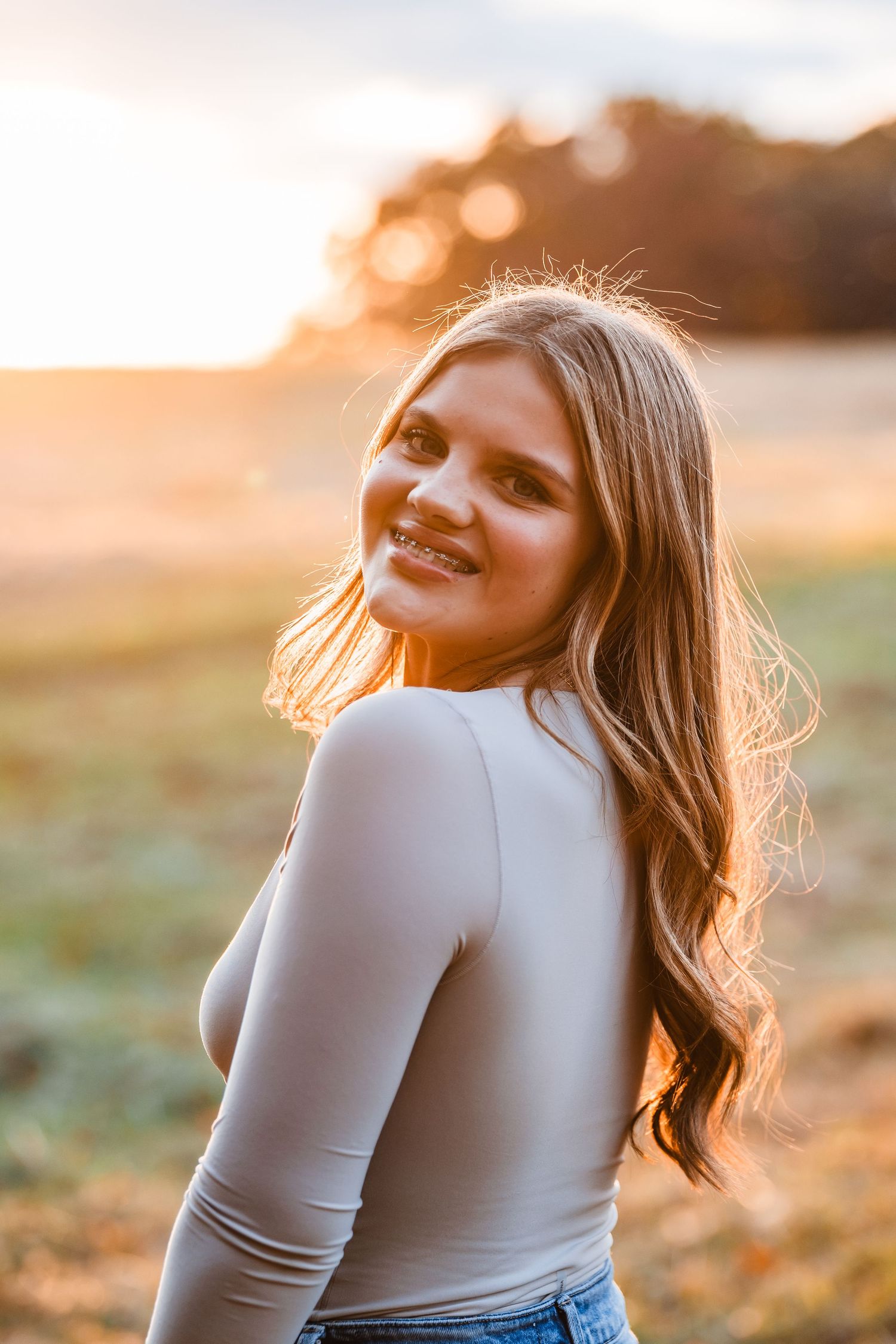 A person in a white long sleeve top poses against a golden sunset background in a field.