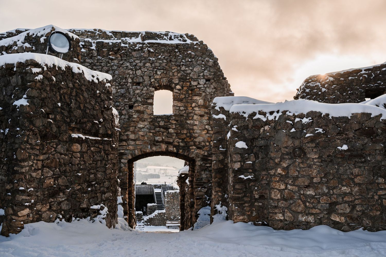 Snow-covered stone archway entrance of an ancient fortress ruin against a moody winter sky.