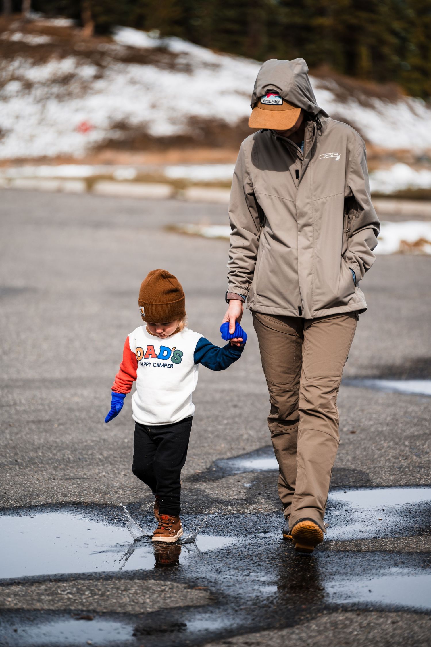 A parent and toddler splash through puddles together on a winter day with patches of snow visible in the background.