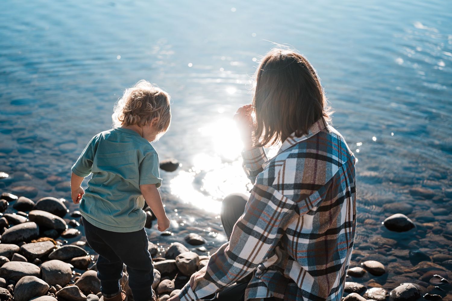 People sitting on a rocky beach shore with sunlight sparkling on bright blue lake waters.