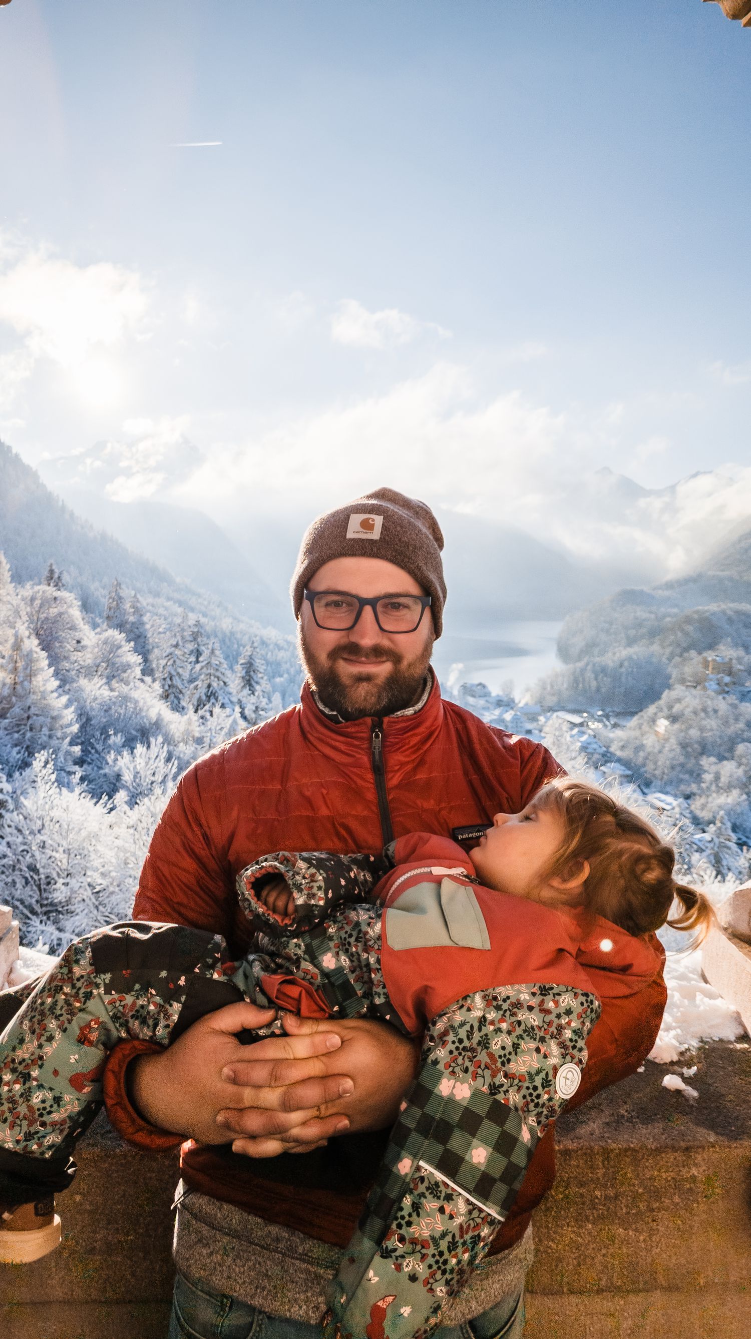 Person in a red jacket relaxes on a stone wall with snowy mountain peaks in the background.