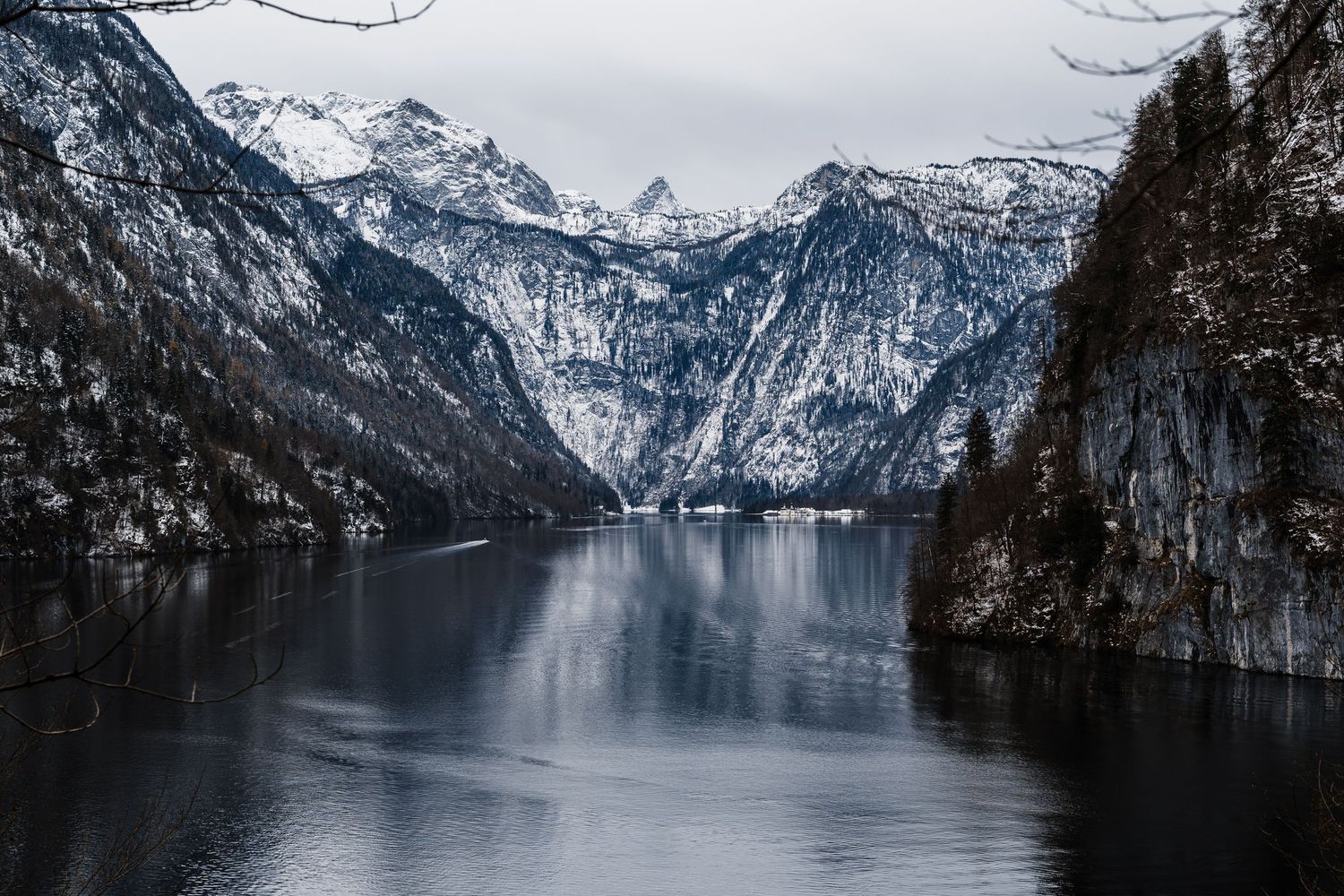 A panoramic view of snow-capped mountains surrounding a serene alpine lake with dark reflective waters and forested shores.