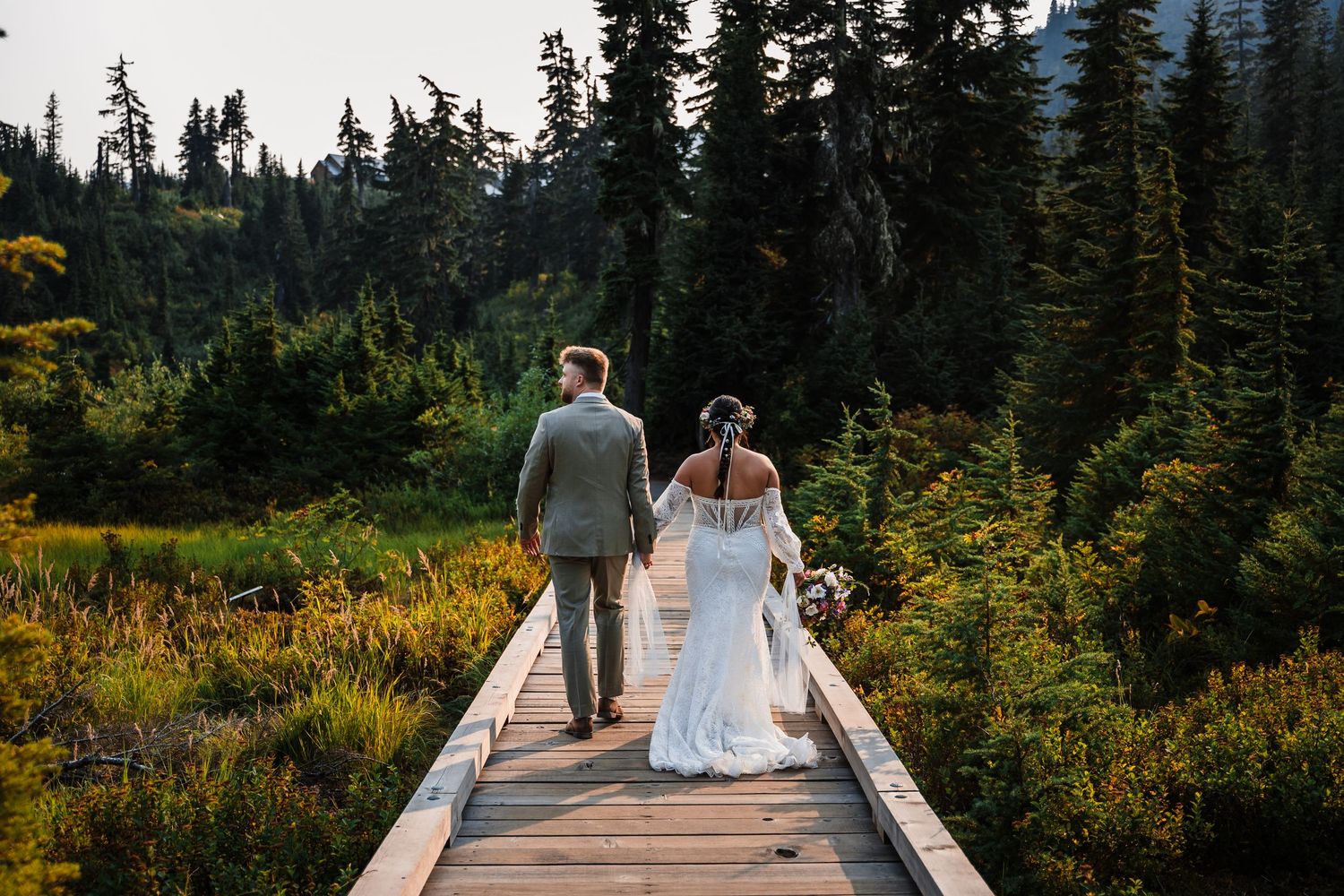 A couple walks along a wooden boardwalk surrounded by evergreen forest and mountain scenery.