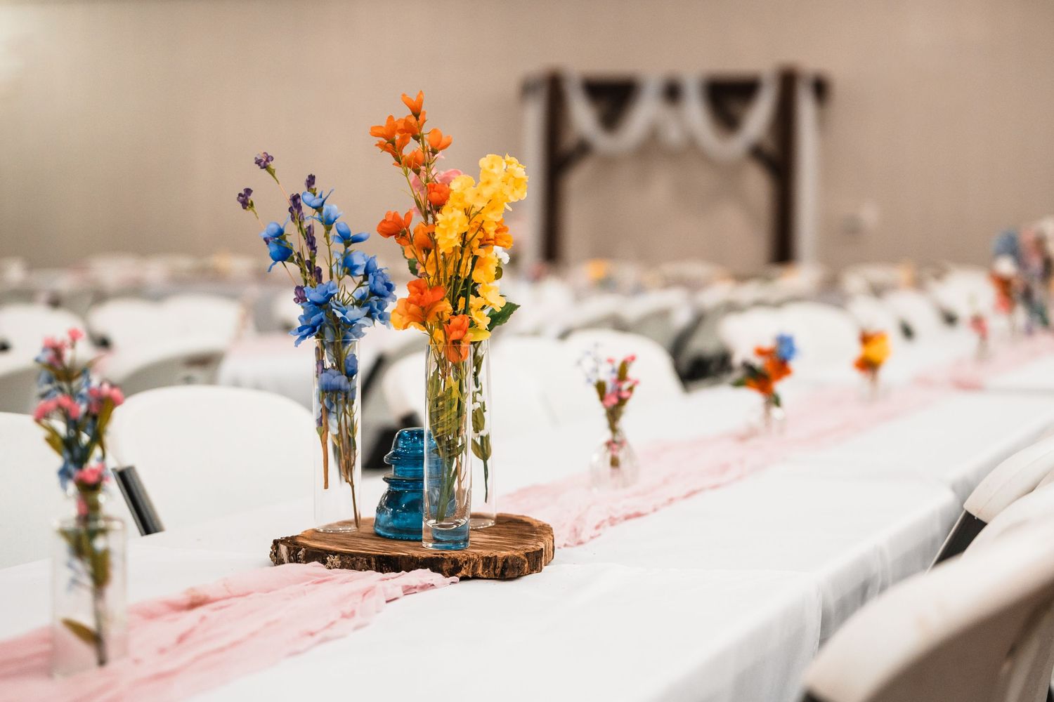 Wedding reception table decorated with colorful wildflower centerpieces on wooden slices and white linens.