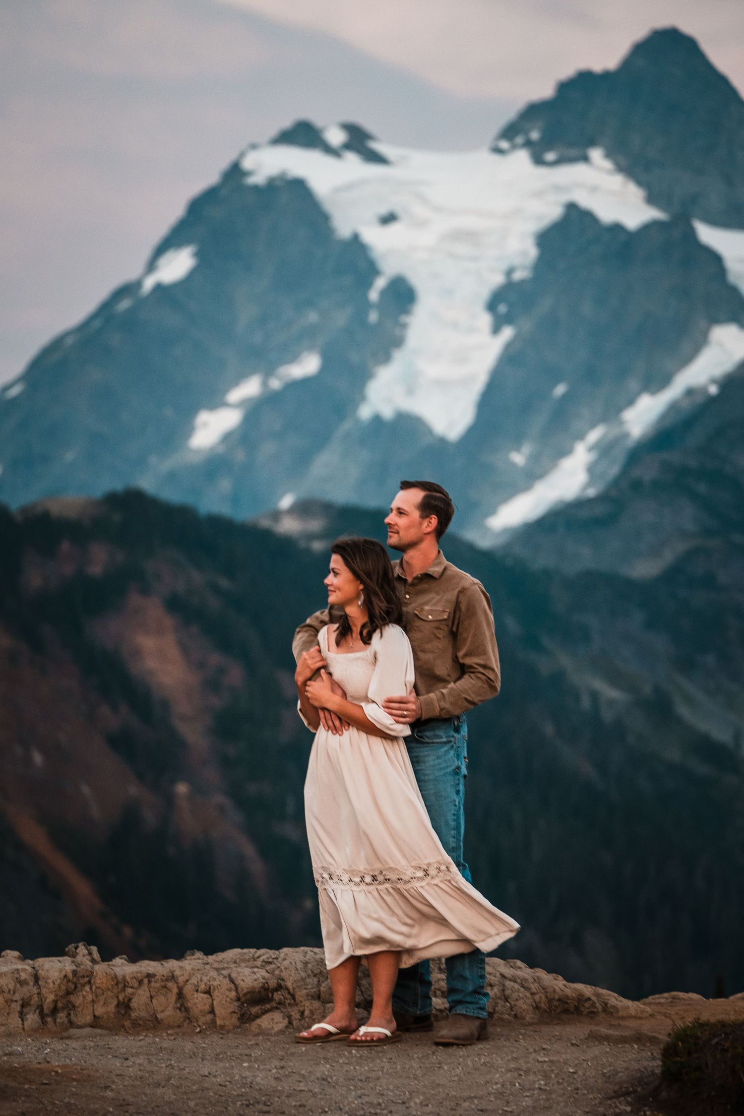 A couple embraces while standing on a cliff edge with a dramatic snow-covered mountain peak in the background.