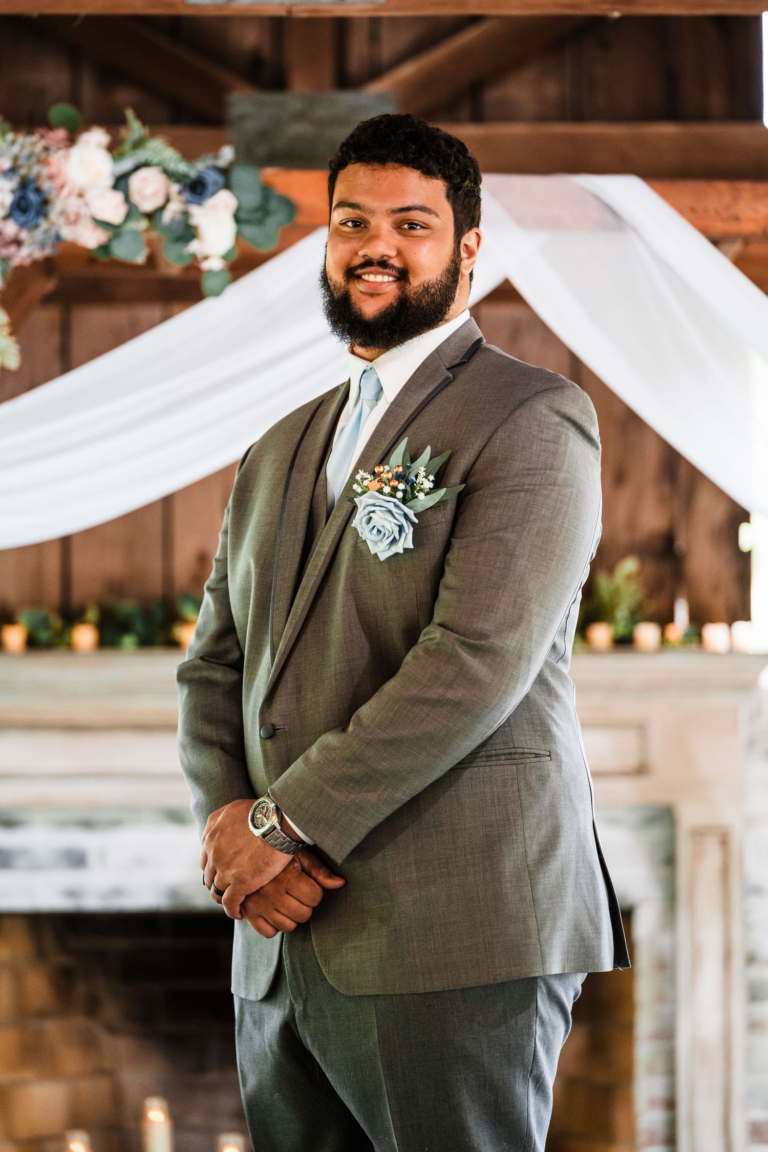 A groom in a gray suit with floral boutonniere stands in front of draped fabric at a rustic venue.