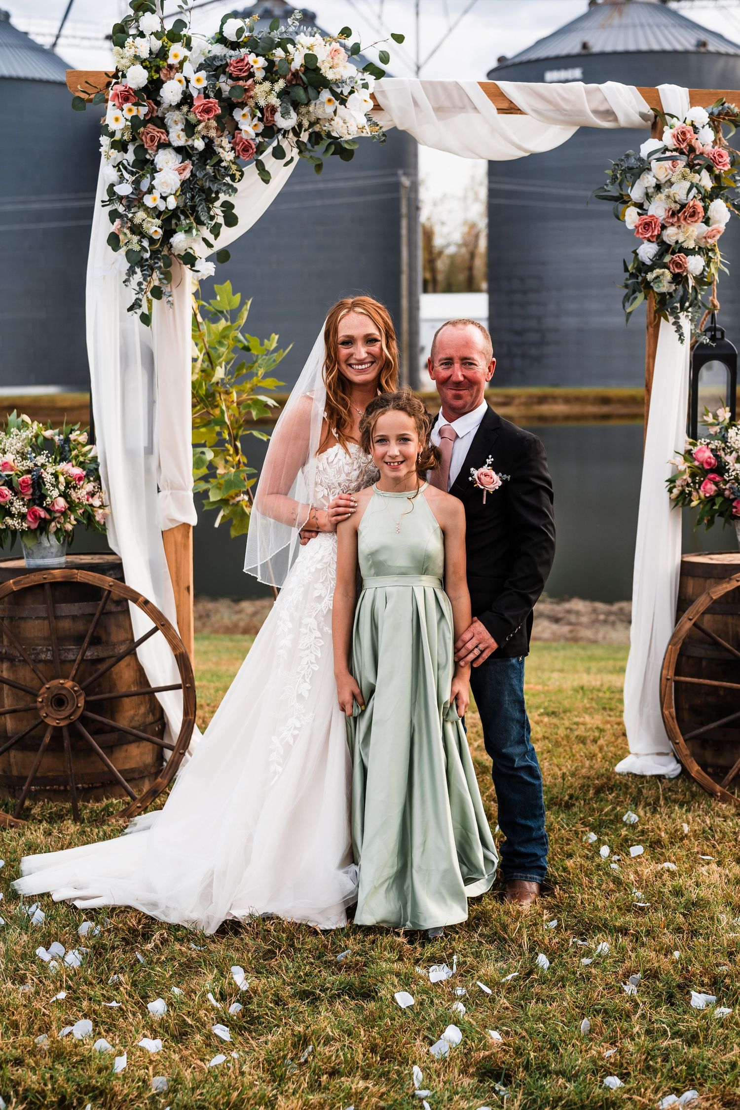 Wedding party posing in front of grain silos with floral arch and rustic decor.