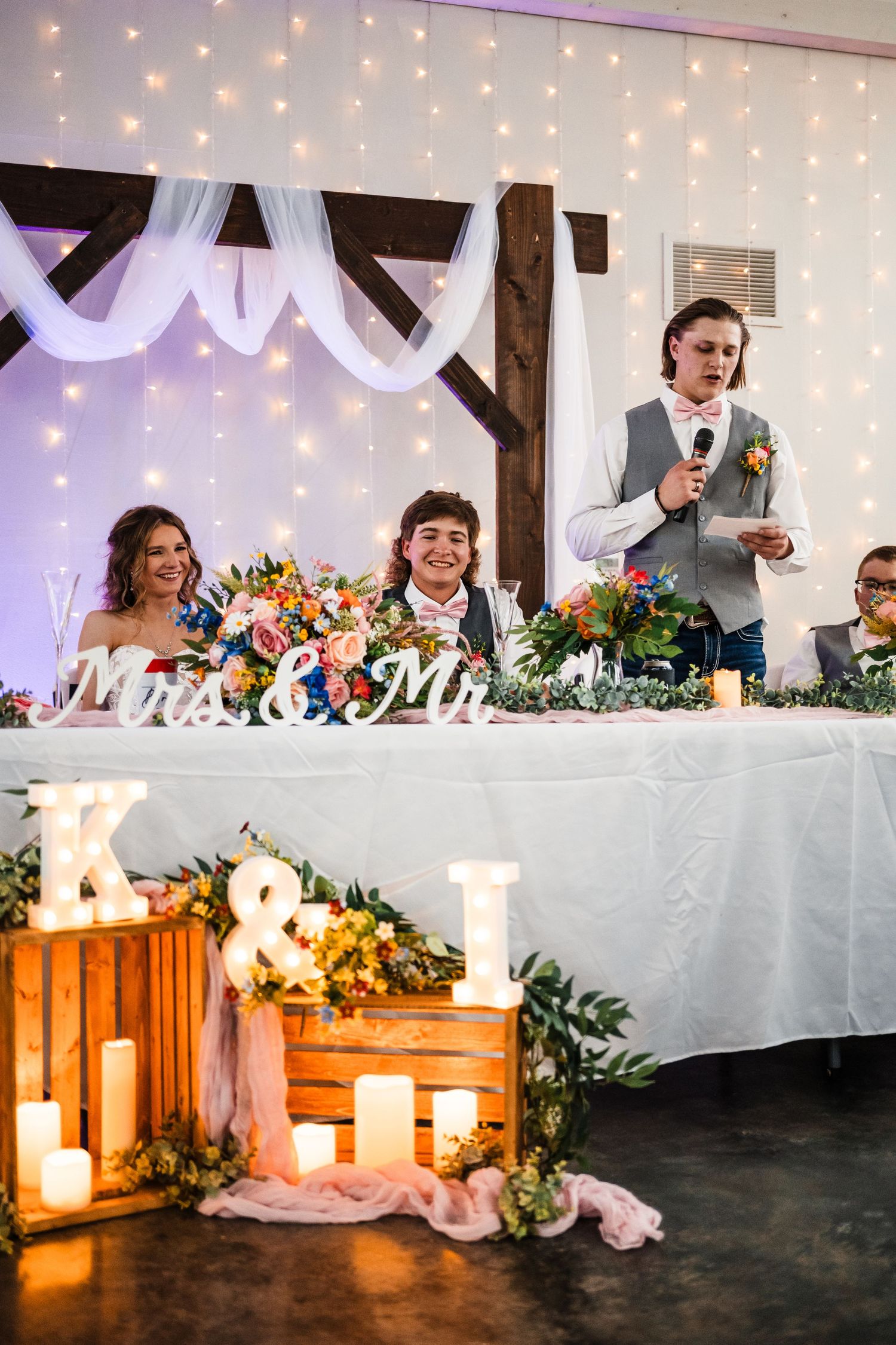 Wedding reception head table with string lights, white draping, and rustic wooden decor.